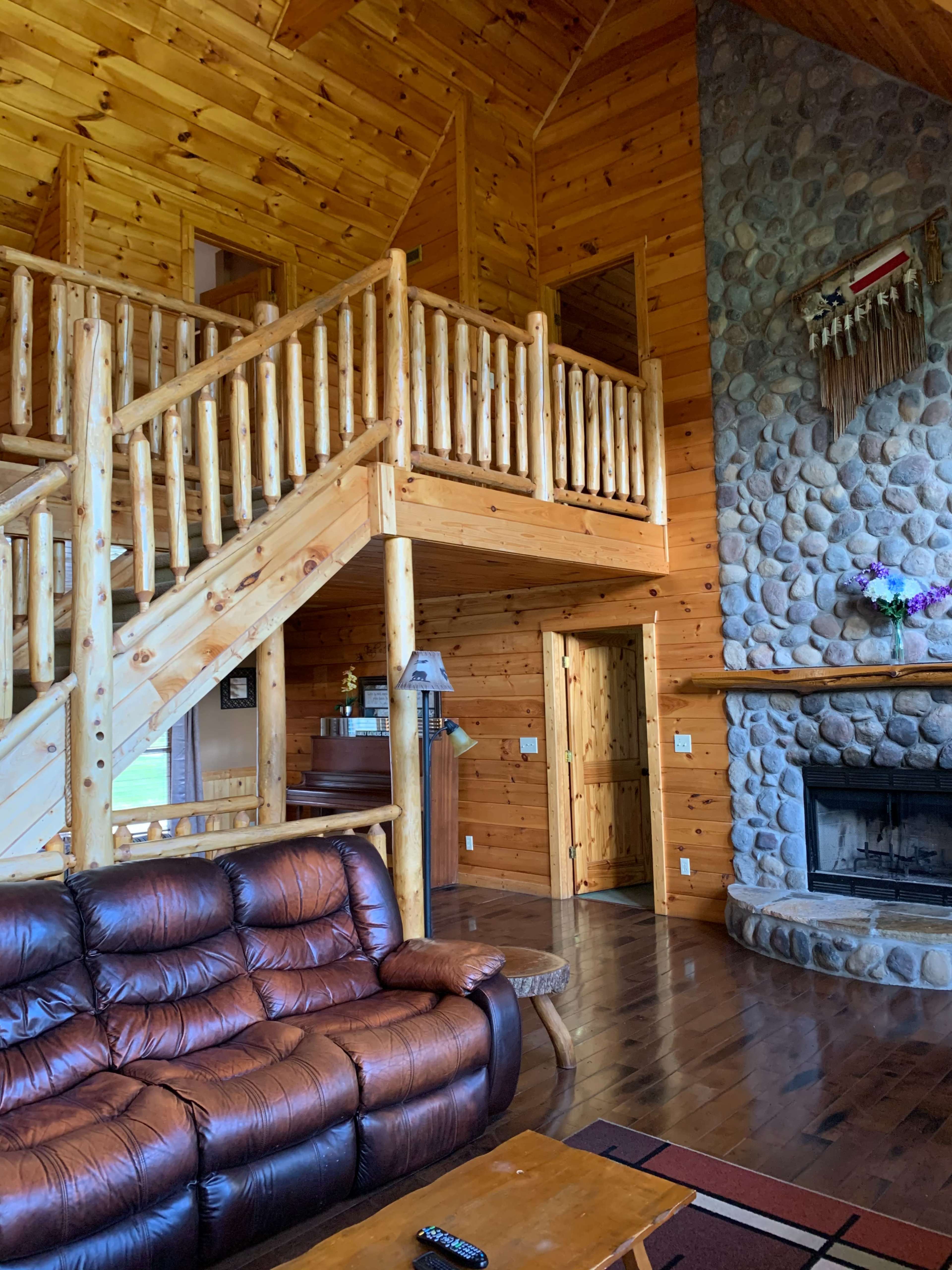 A cozy wooden interior of a cabin with a staircase, stone fireplace, and a leather couch.