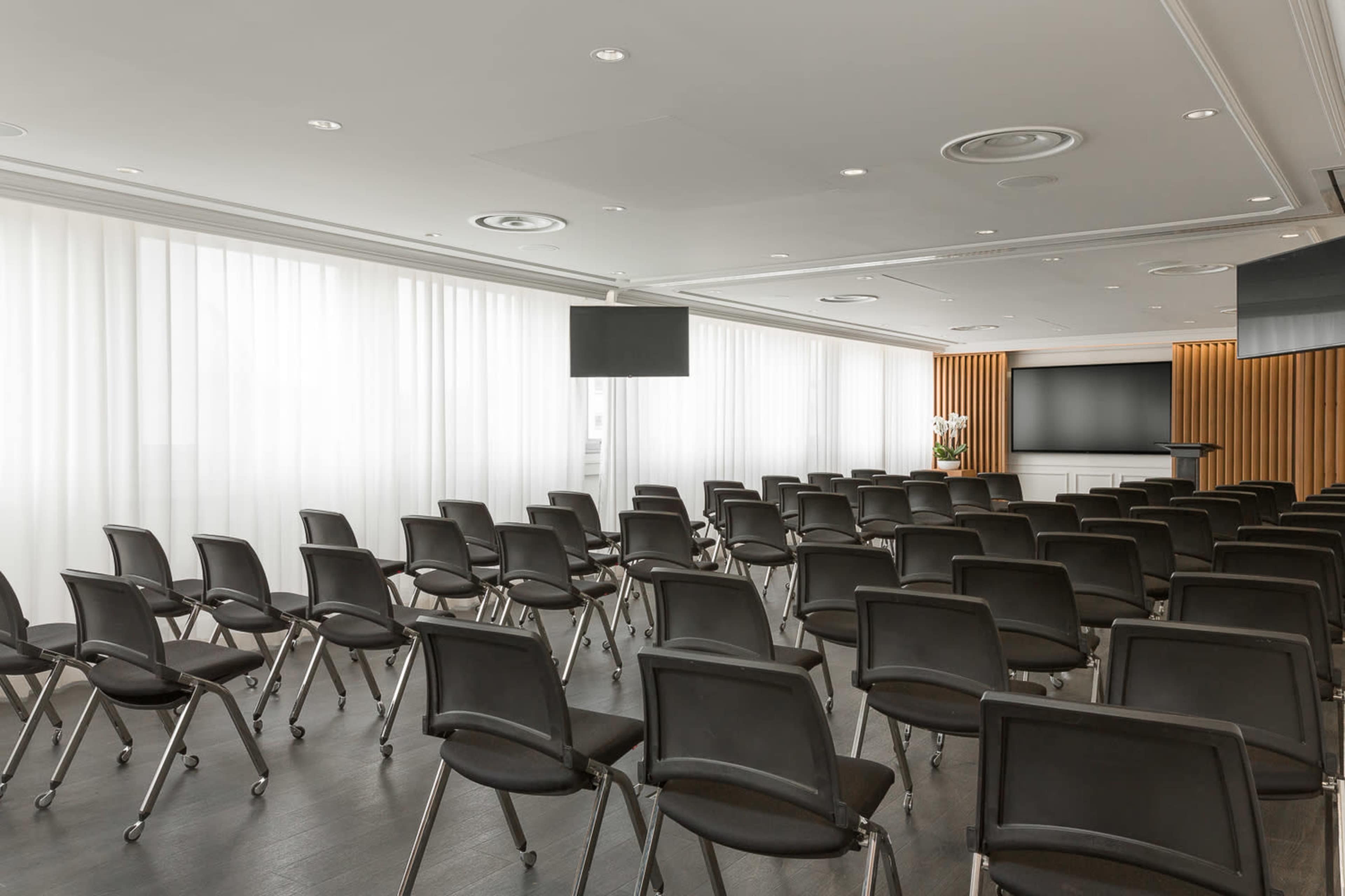 A conference room is set up with rows of black chairs facing a large screen.