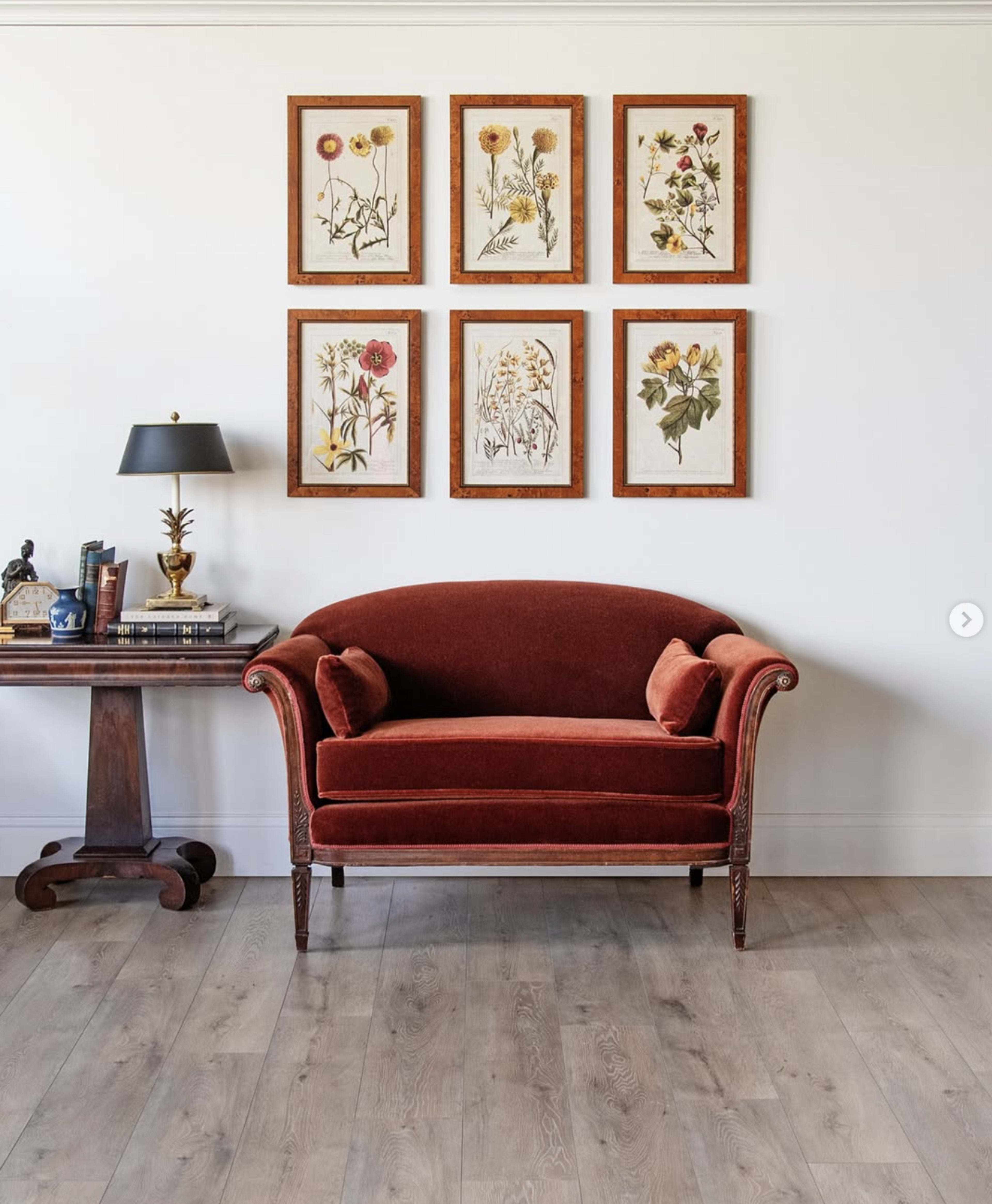 A red velvet loveseat is placed against a wall adorned with framed botanical prints, next to a wooden side table with a lamp and books.