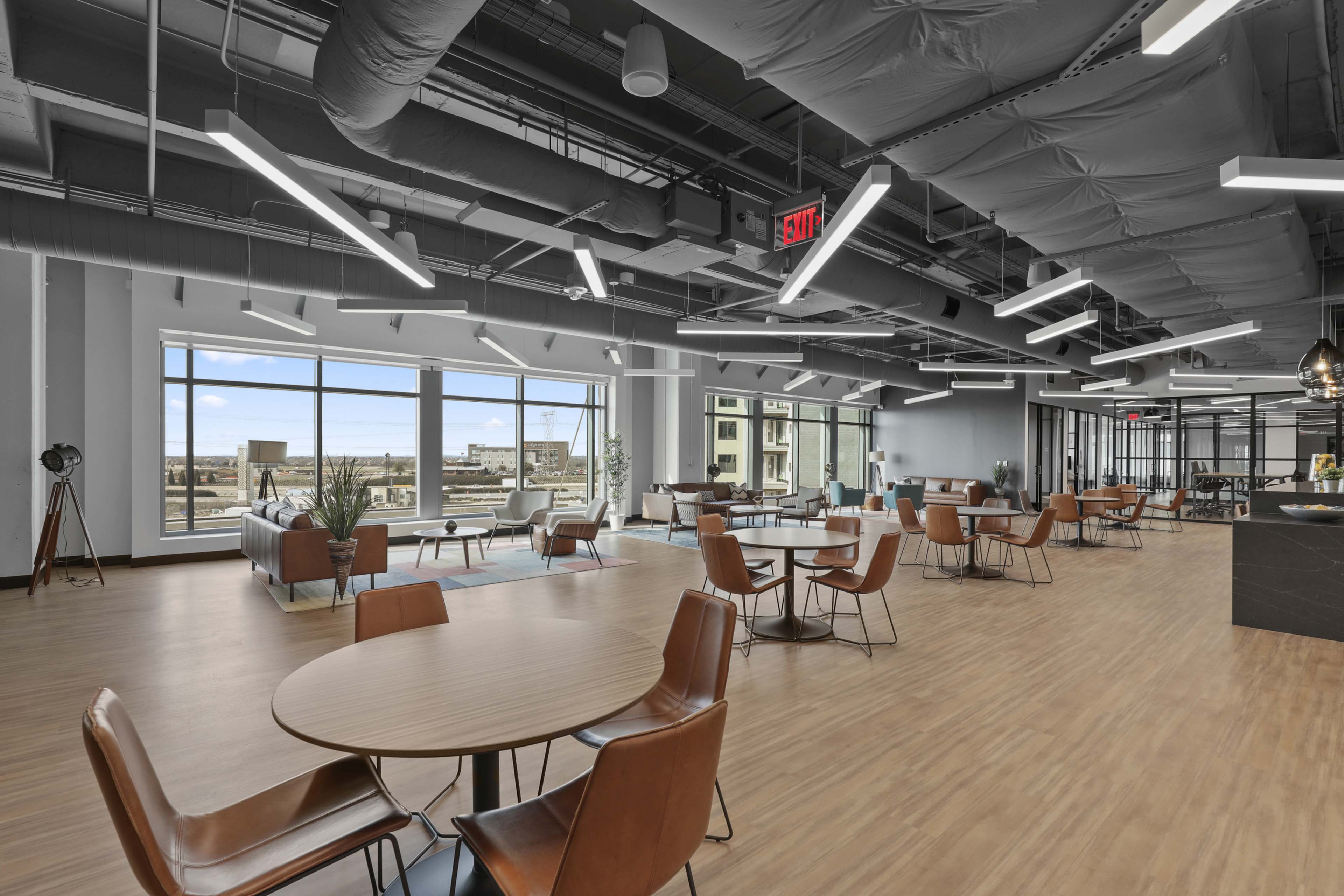 A modern office lounge area with wooden tables and chairs, large windows, and a spacious layout.