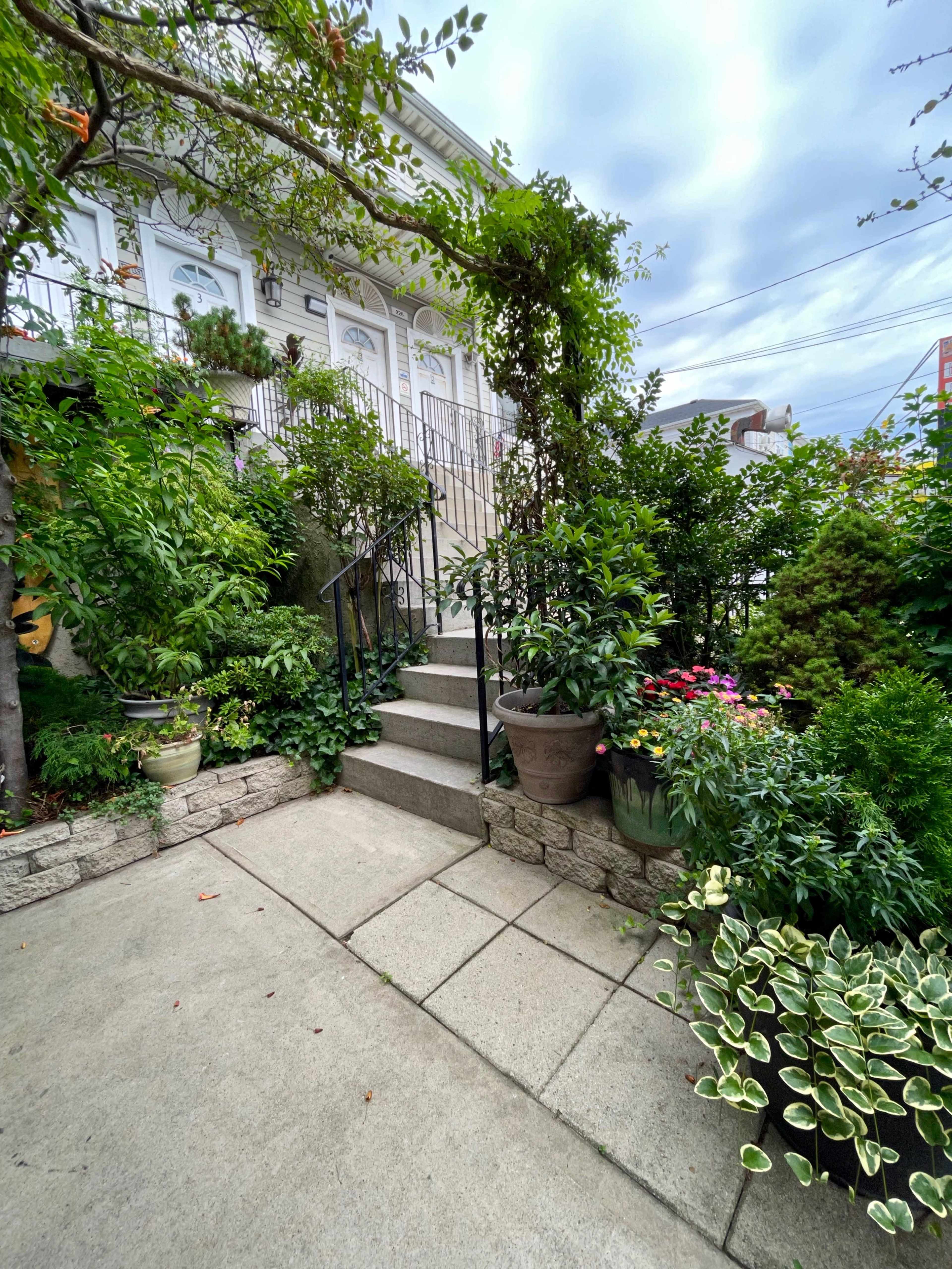 A stone pathway leads to a set of stairs flanked by various potted plants and greenery.