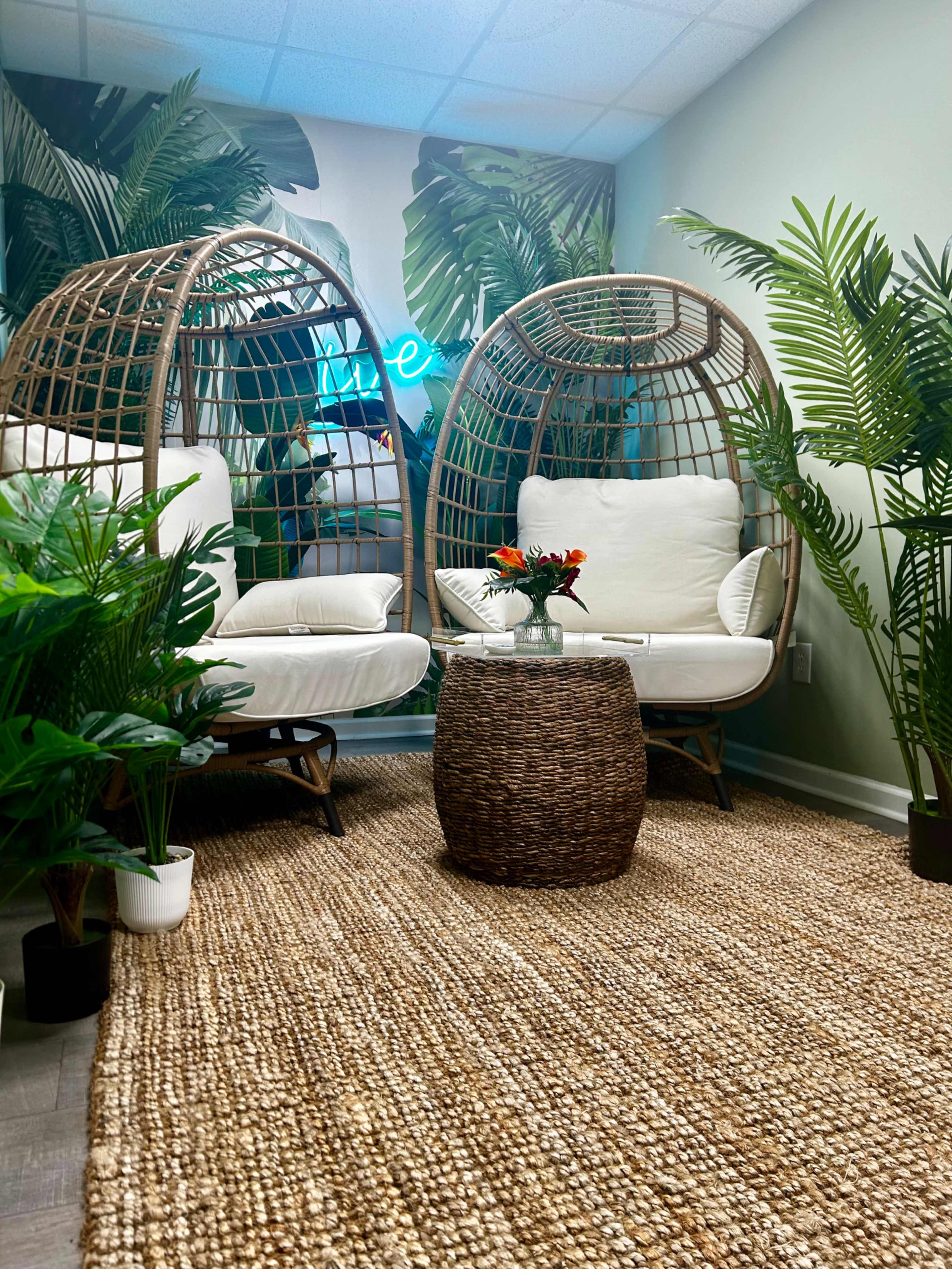 The image shows two large, round wicker chairs placed in a corner, surrounded by greenery and a woven rug on the floor.