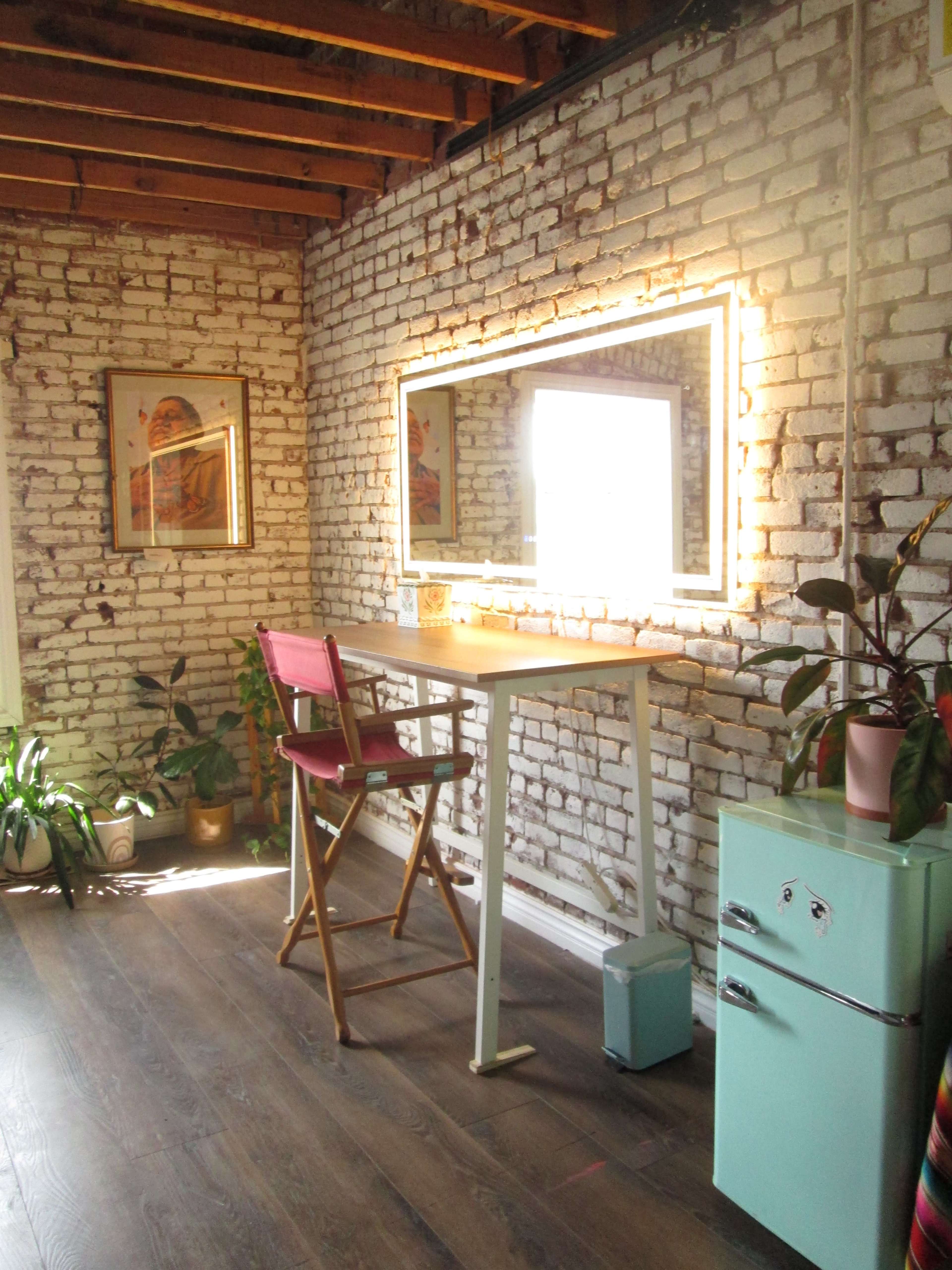 The image shows a well-lit room featuring a white brick wall, a small wooden table with a mirror framed in neon lighting, a pink chair, and a vintage mint green mini fridge.