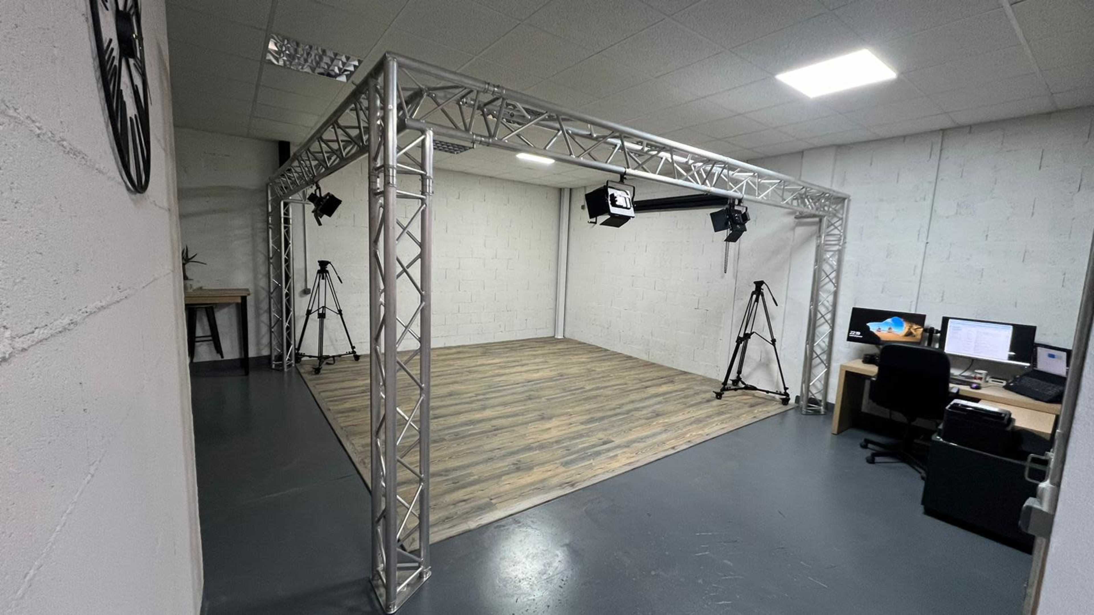 A minimalist studio space with a wooden floor, surrounded by a metal truss frame and equipped with lighting and camera tripods.
