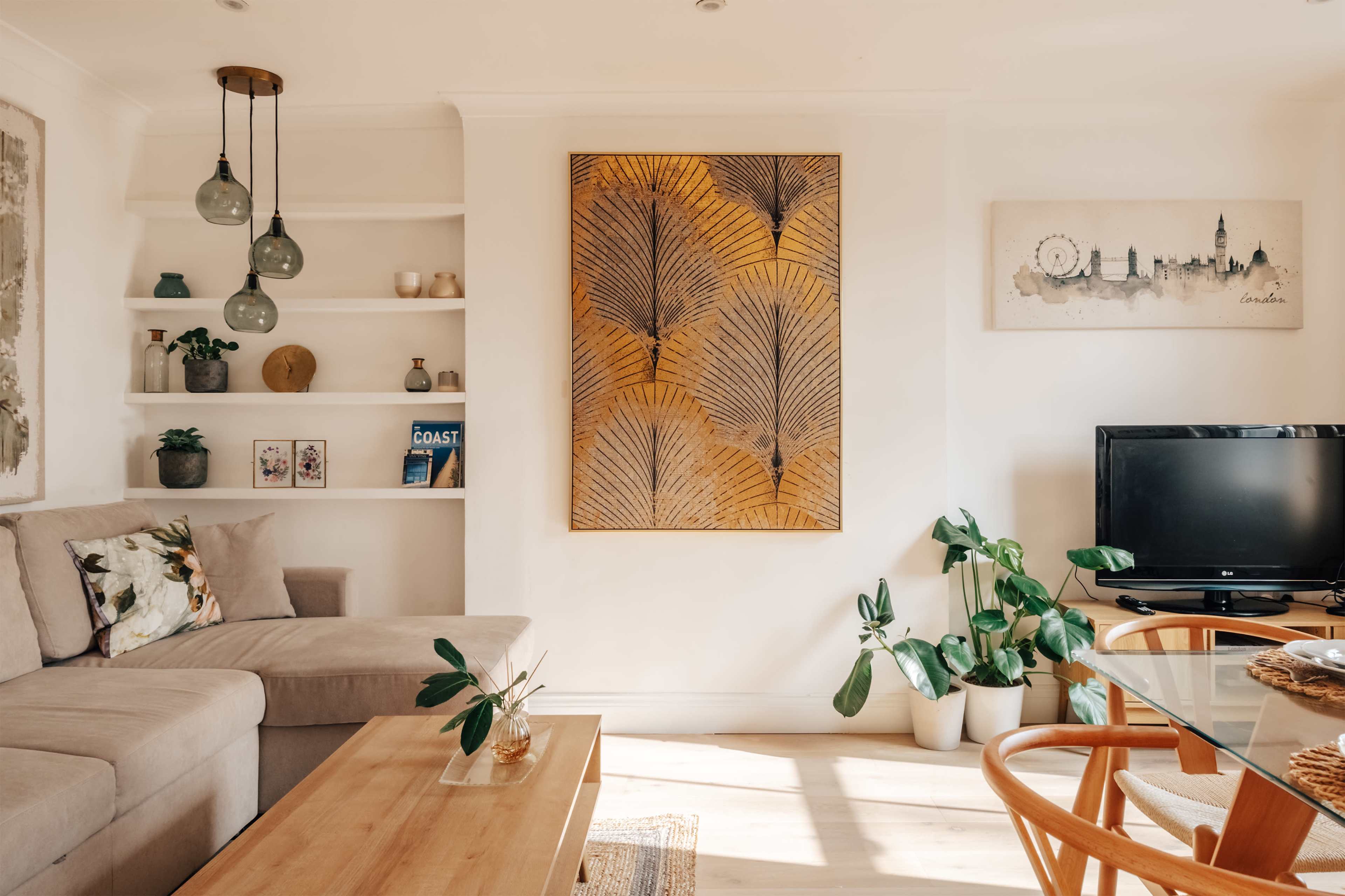 The image shows a bright living room with a corner couch, a wooden coffee table, a flat-screen TV, and decorative wall art.