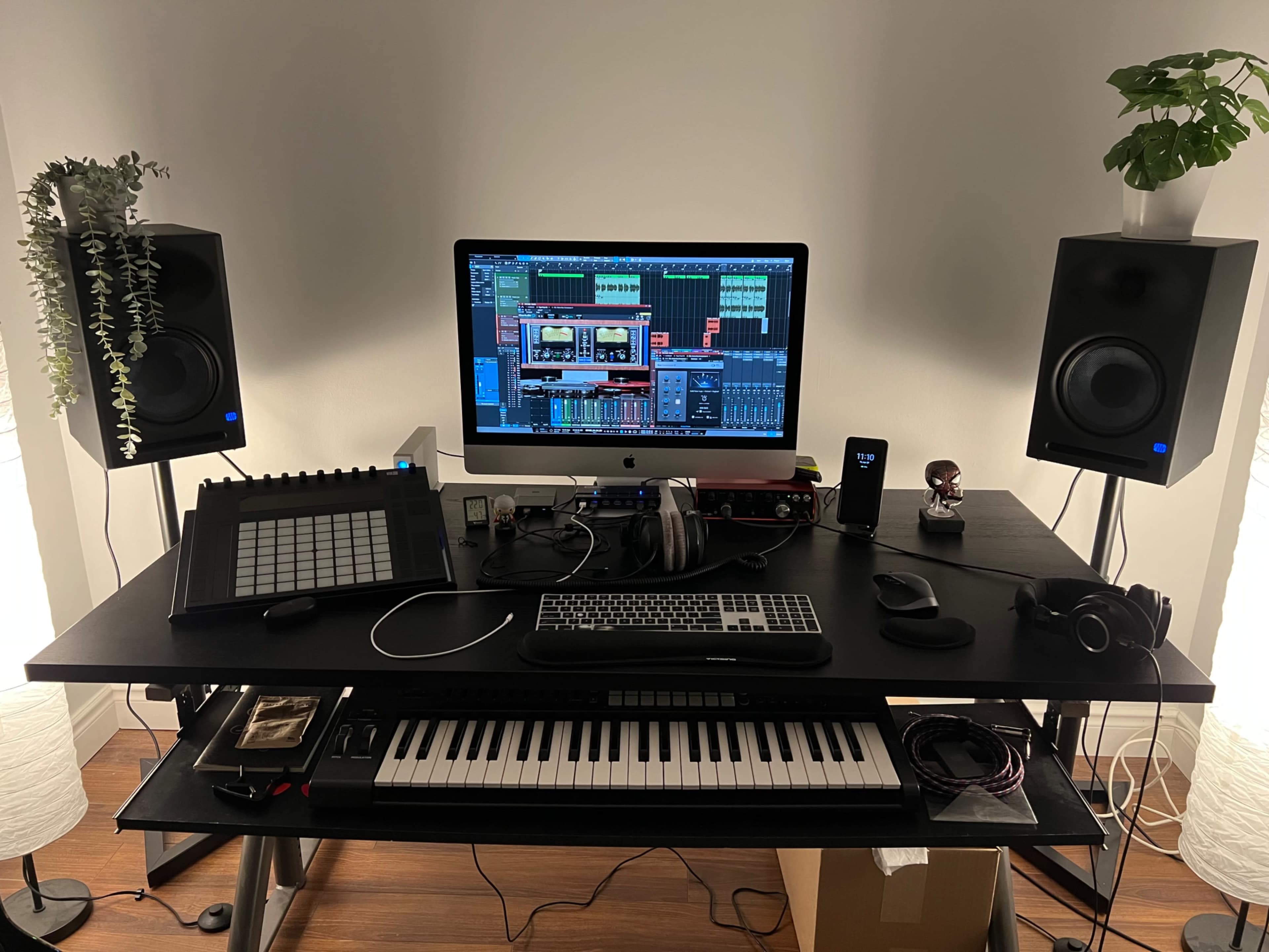 A music production setup featuring a computer monitor, audio speakers, MIDI keyboard, a drum pad controller, headphones, and various cables on a black desk.