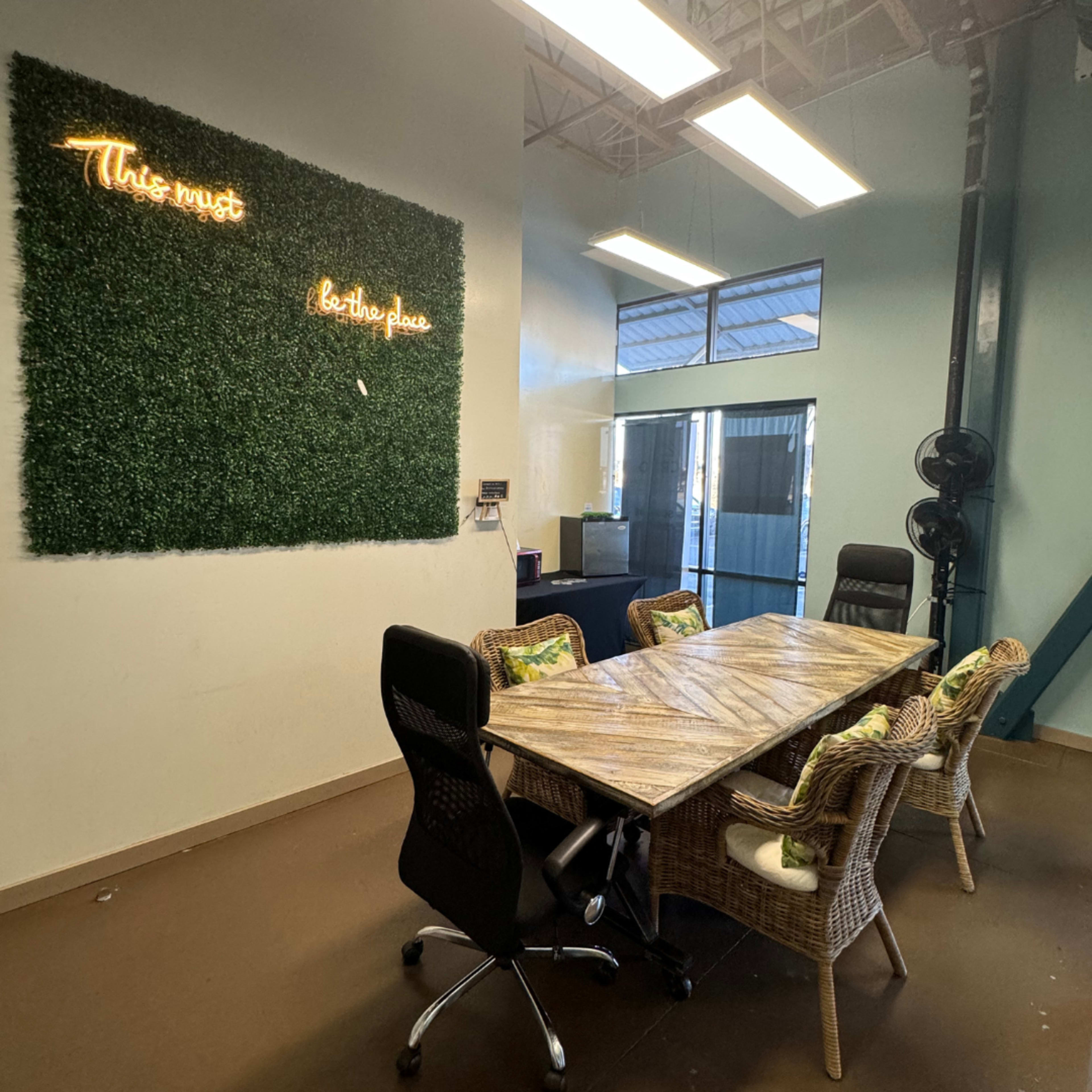 A modern meeting room features a wooden table surrounded by black and rattan chairs, with a green wall displaying neon text.