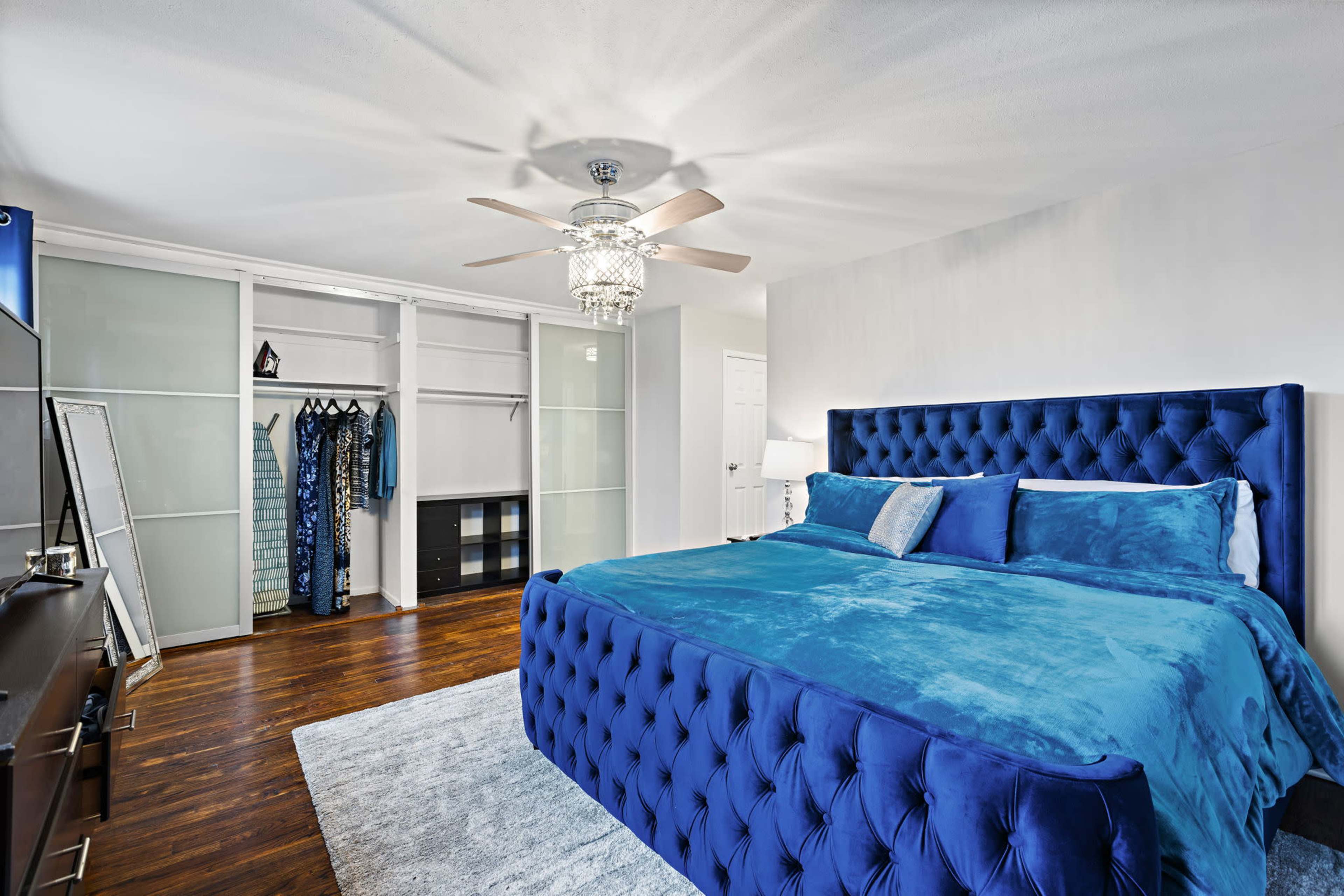 A spacious bedroom features a large blue tufted bed, a stylish ceiling fan, and an organized closet with hanging clothes.