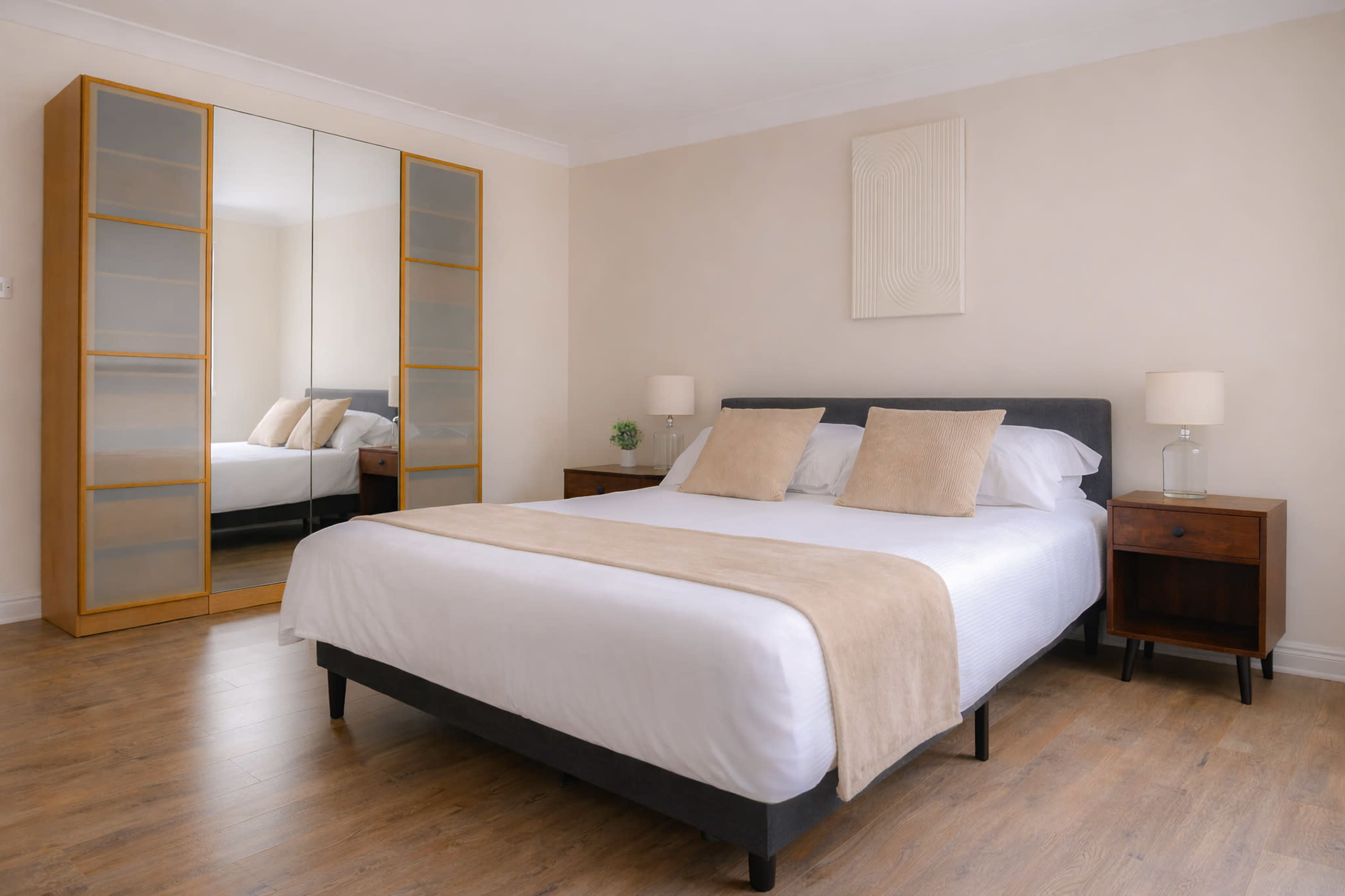 The image shows a well-furnished bedroom featuring a large bed with white linens, two bedside tables with lamps, and a mirrored wardrobe against the wall.