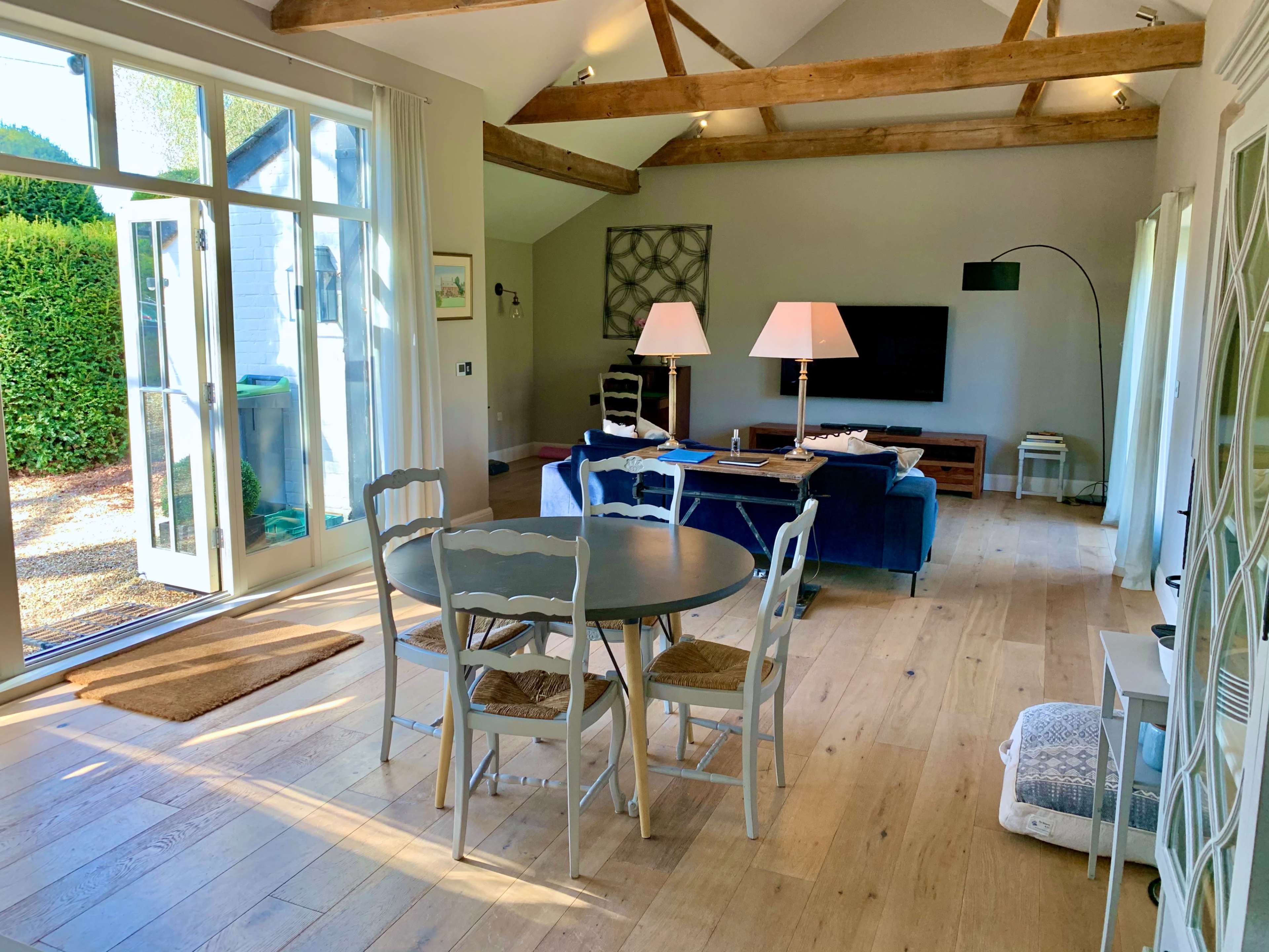 A bright, modern living space features a round dining table, two large lamps, and a sectional sofa with large windows and open doors leading to a garden.