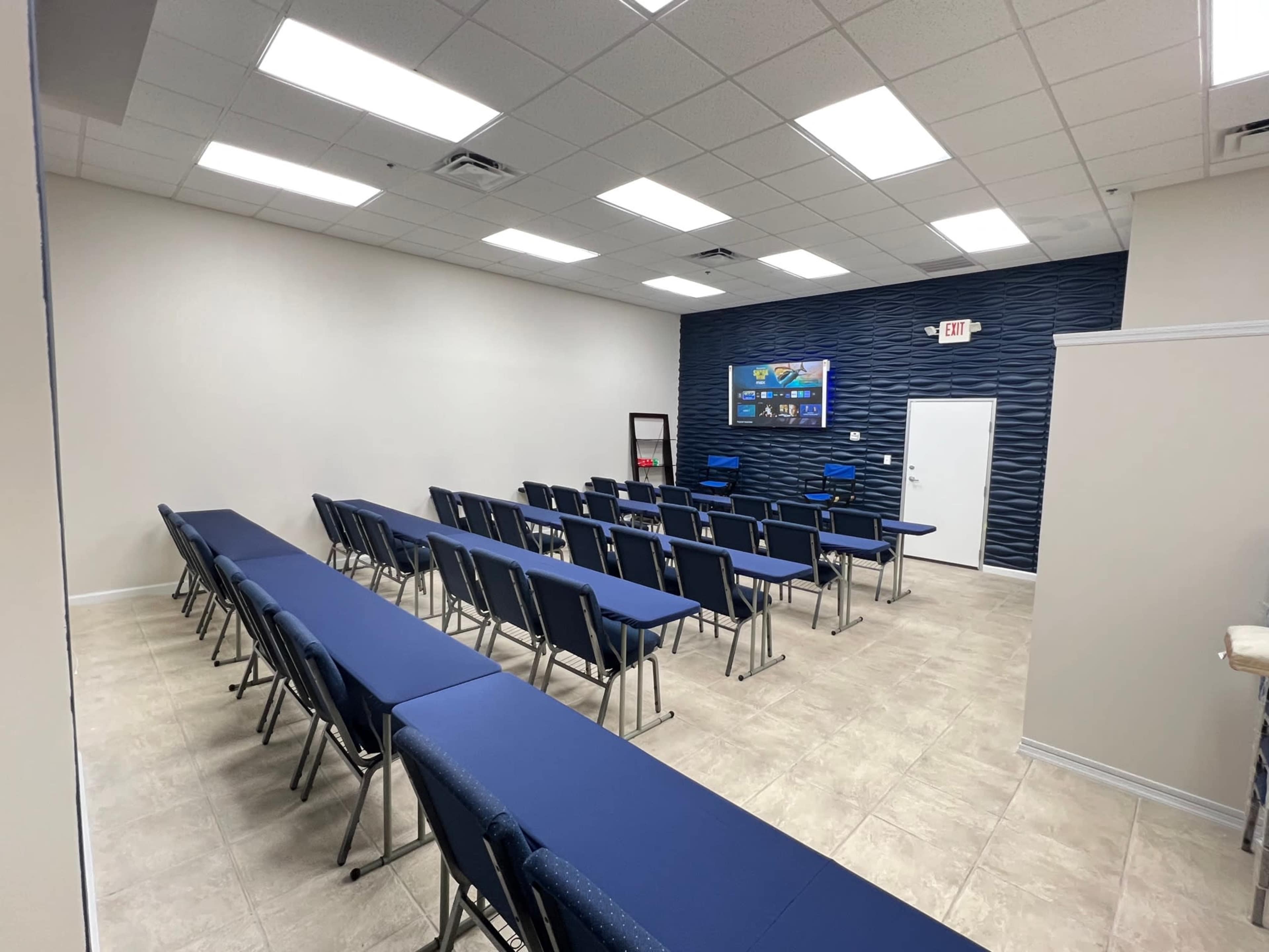 A classroom or conference room with rows of blue chairs and a wall-mounted television at the front.