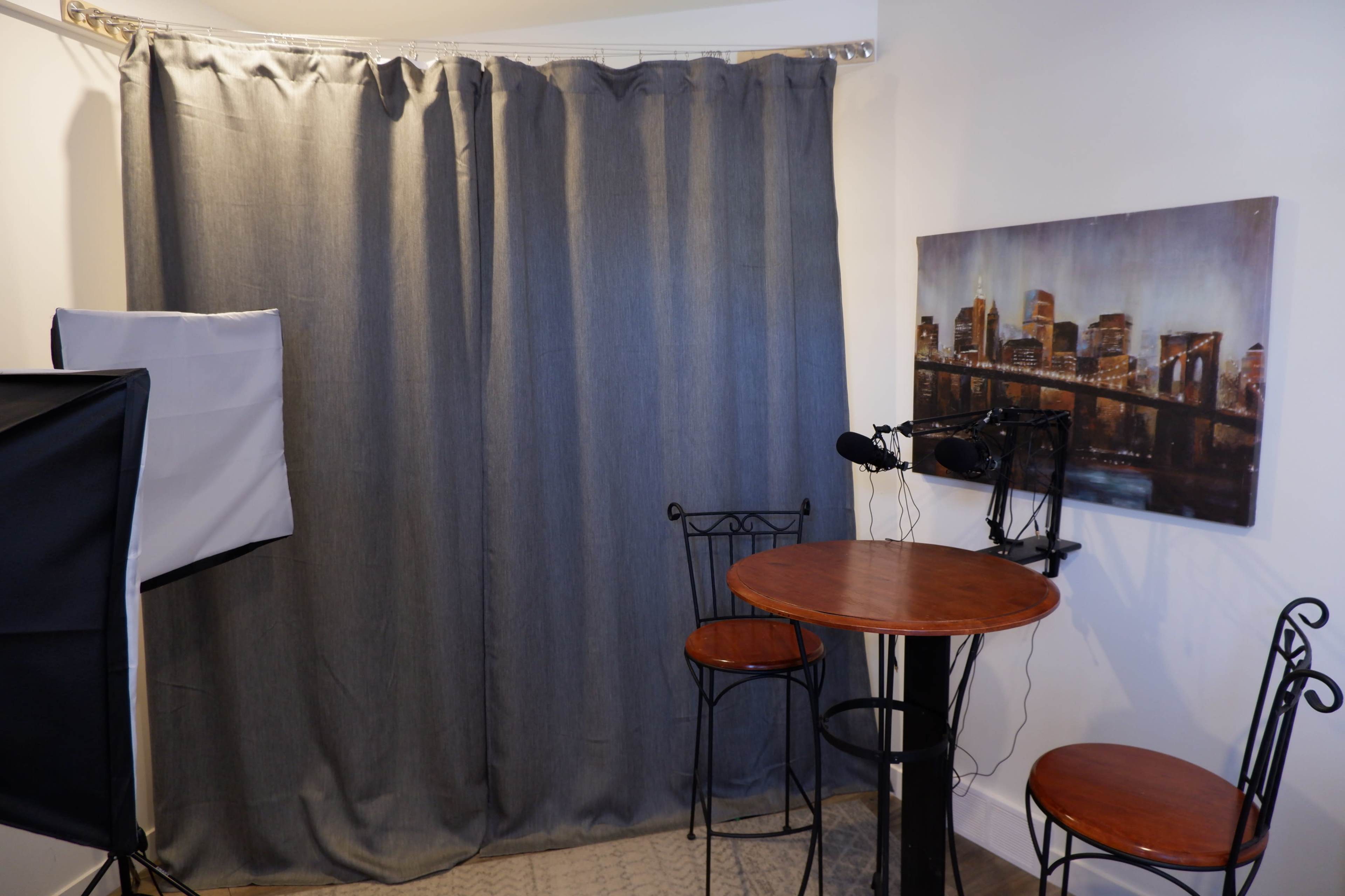 The image shows a small studio space with a round table and two metal chairs, a microphone setup, and a backdrop of gray curtains.