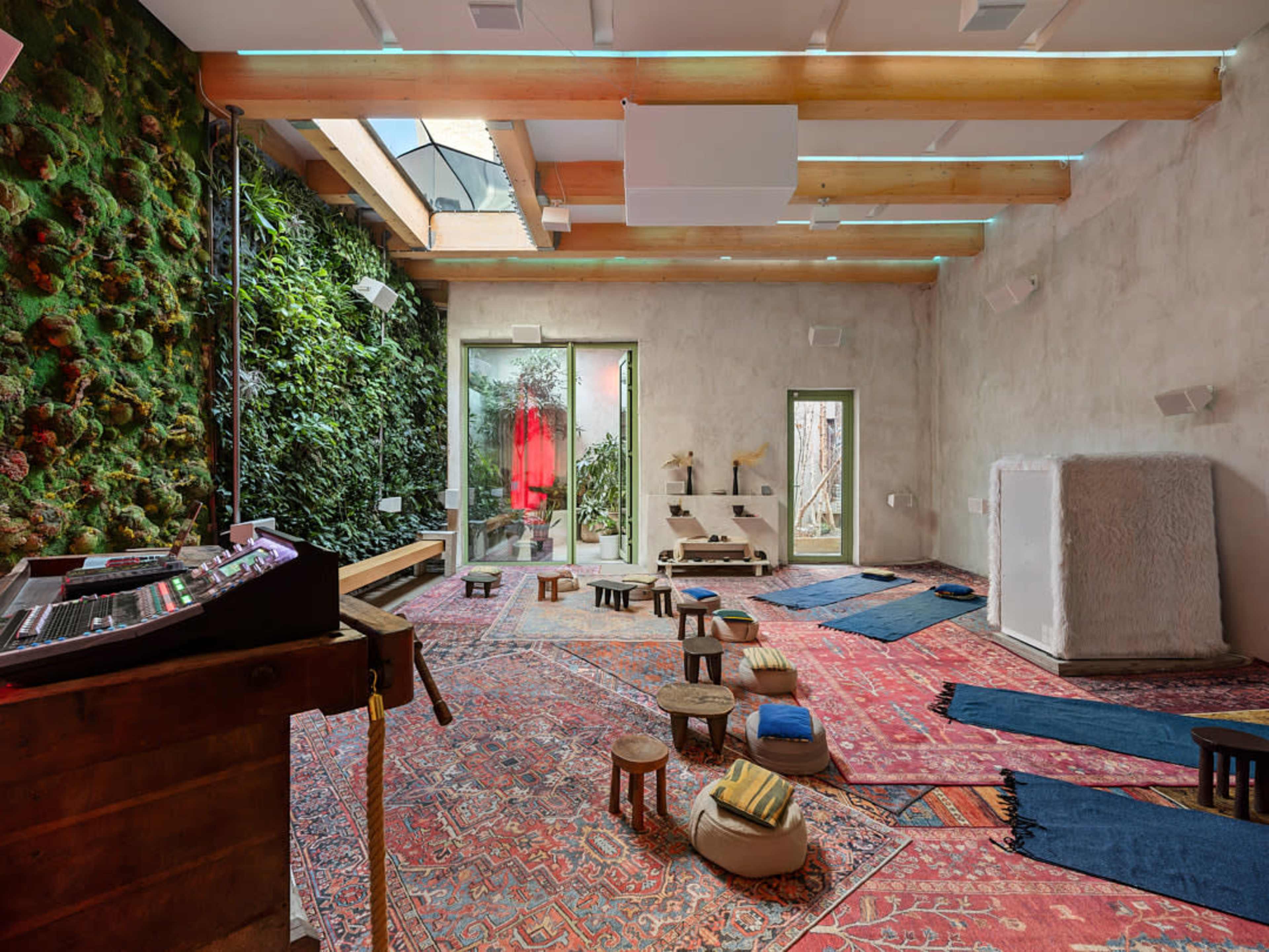 The image shows a spacious, brightly lit room featuring a mix of wooden and textile seating, colorful rugs, a green moss wall, and large windows that open to an outdoor area.