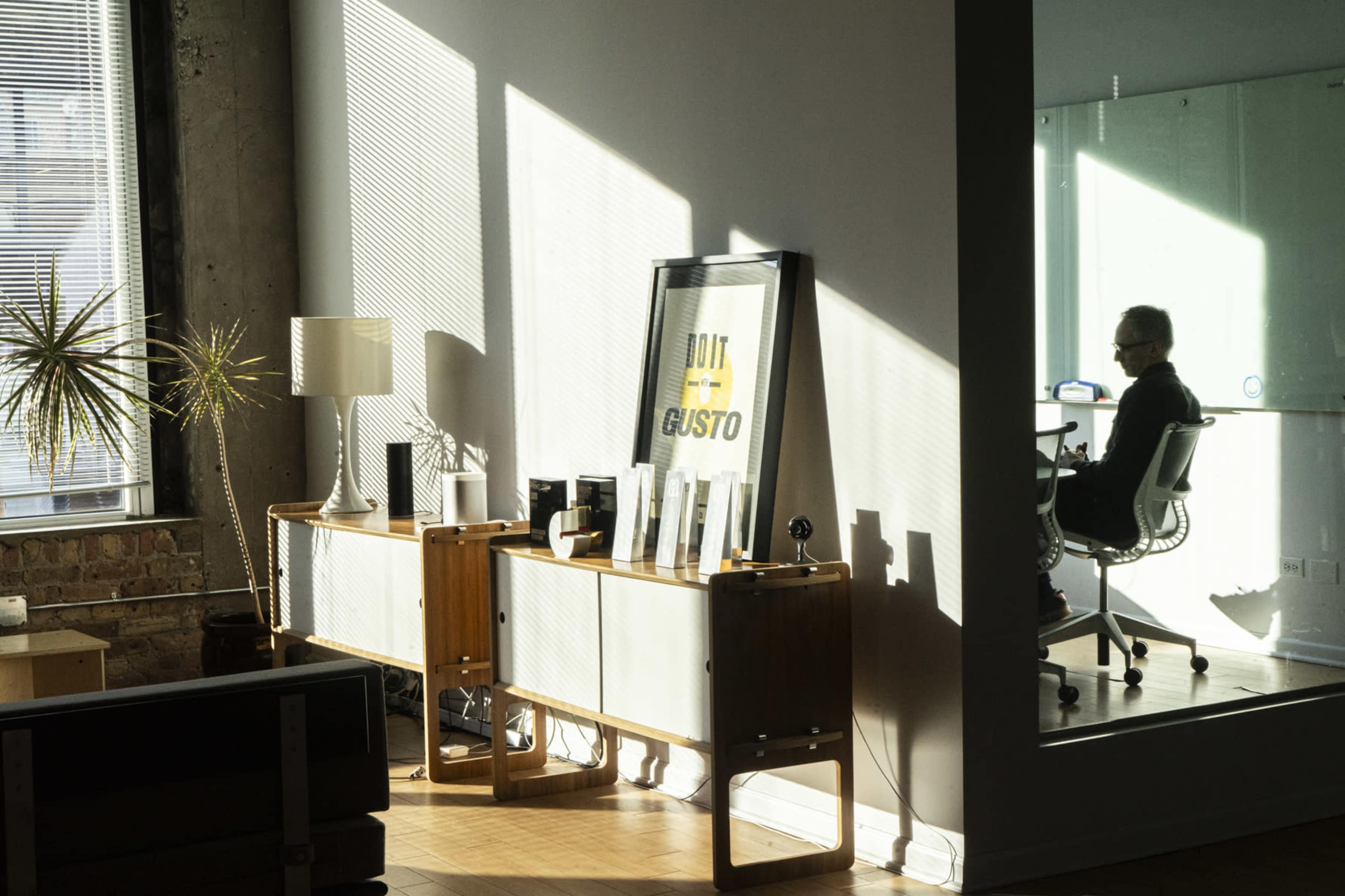 A modern office space features a wooden console table with decorative items and a large framed poster, illuminated by sunlight filtering through the windows, while a person sits at a desk in a glass-walled room.
