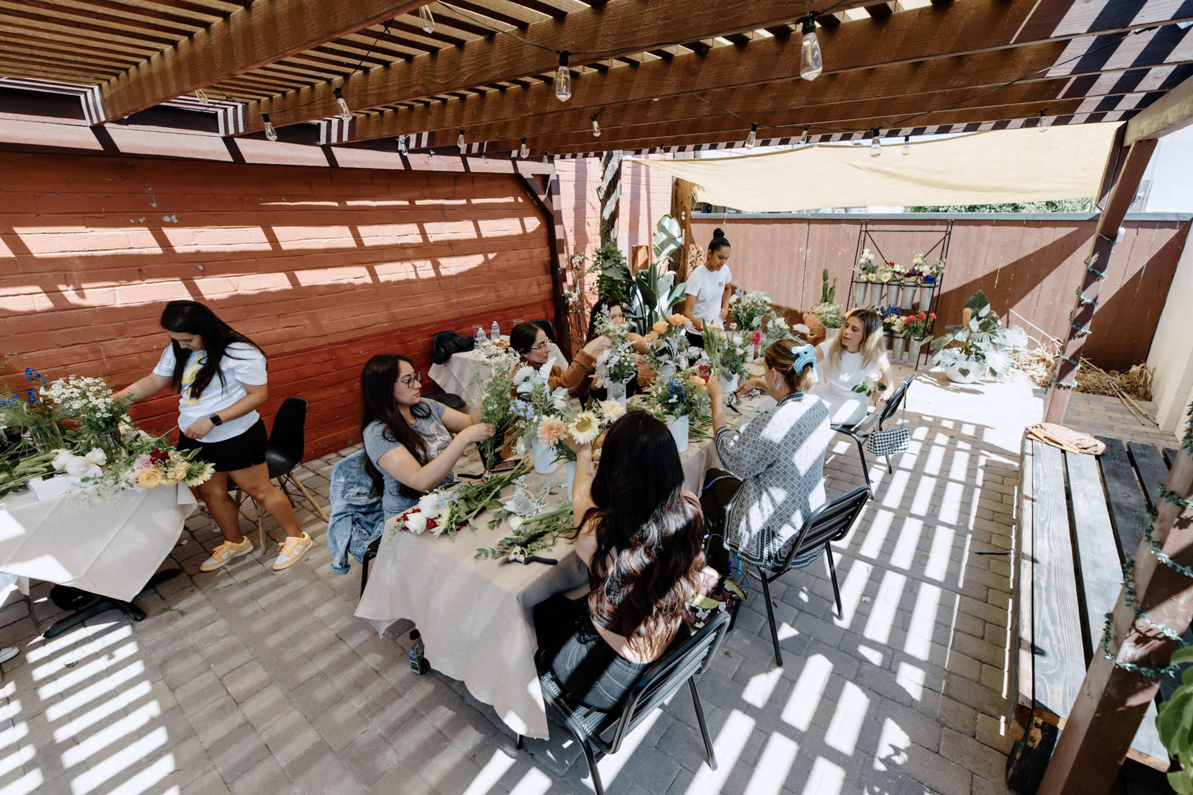 A group of people is gathered around tables in a shaded outdoor space, arranging flowers for a craft activity.