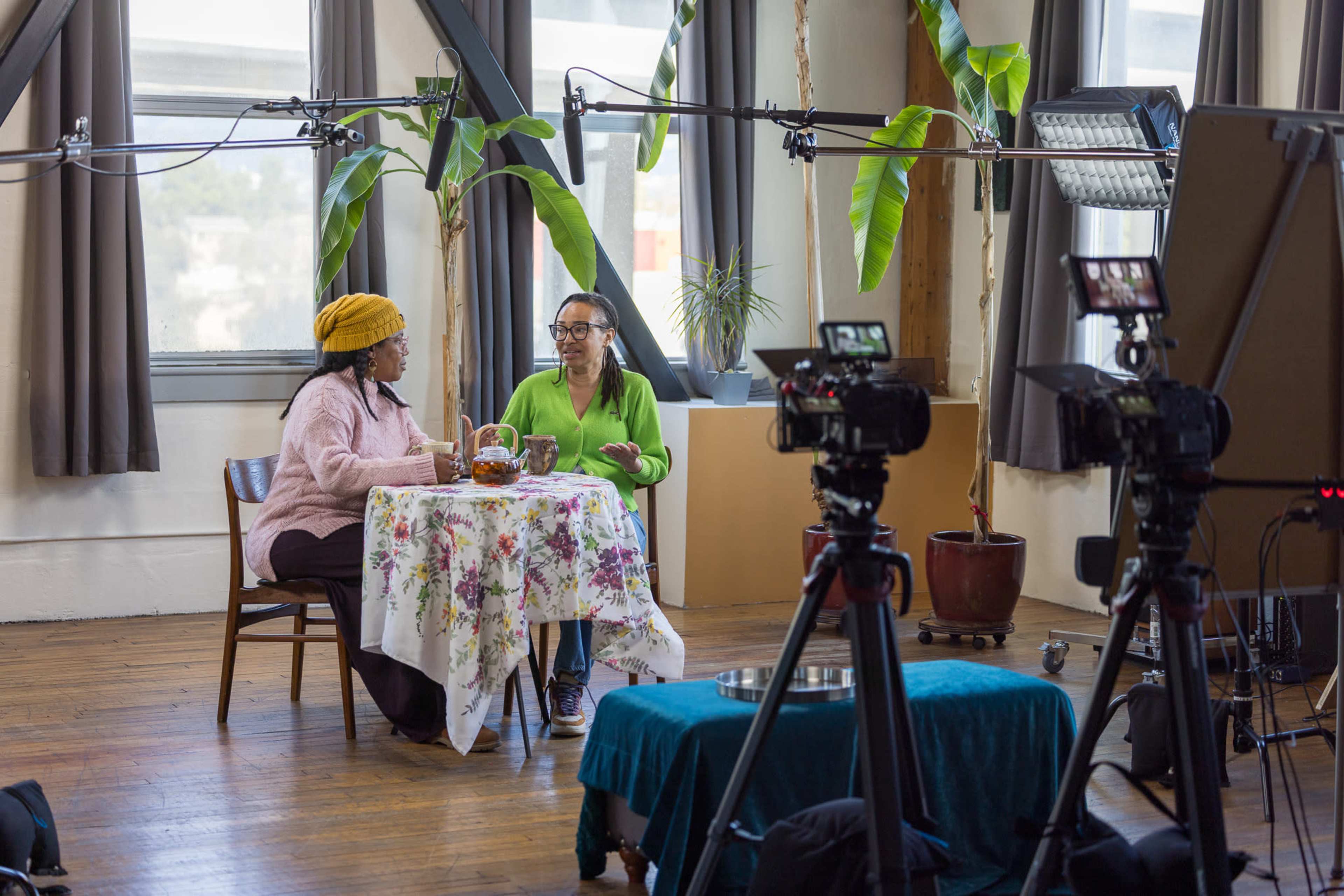 Two women are seated at a table with a floral cloth in a well-lit room, while film cameras and lighting equipment are positioned nearby.