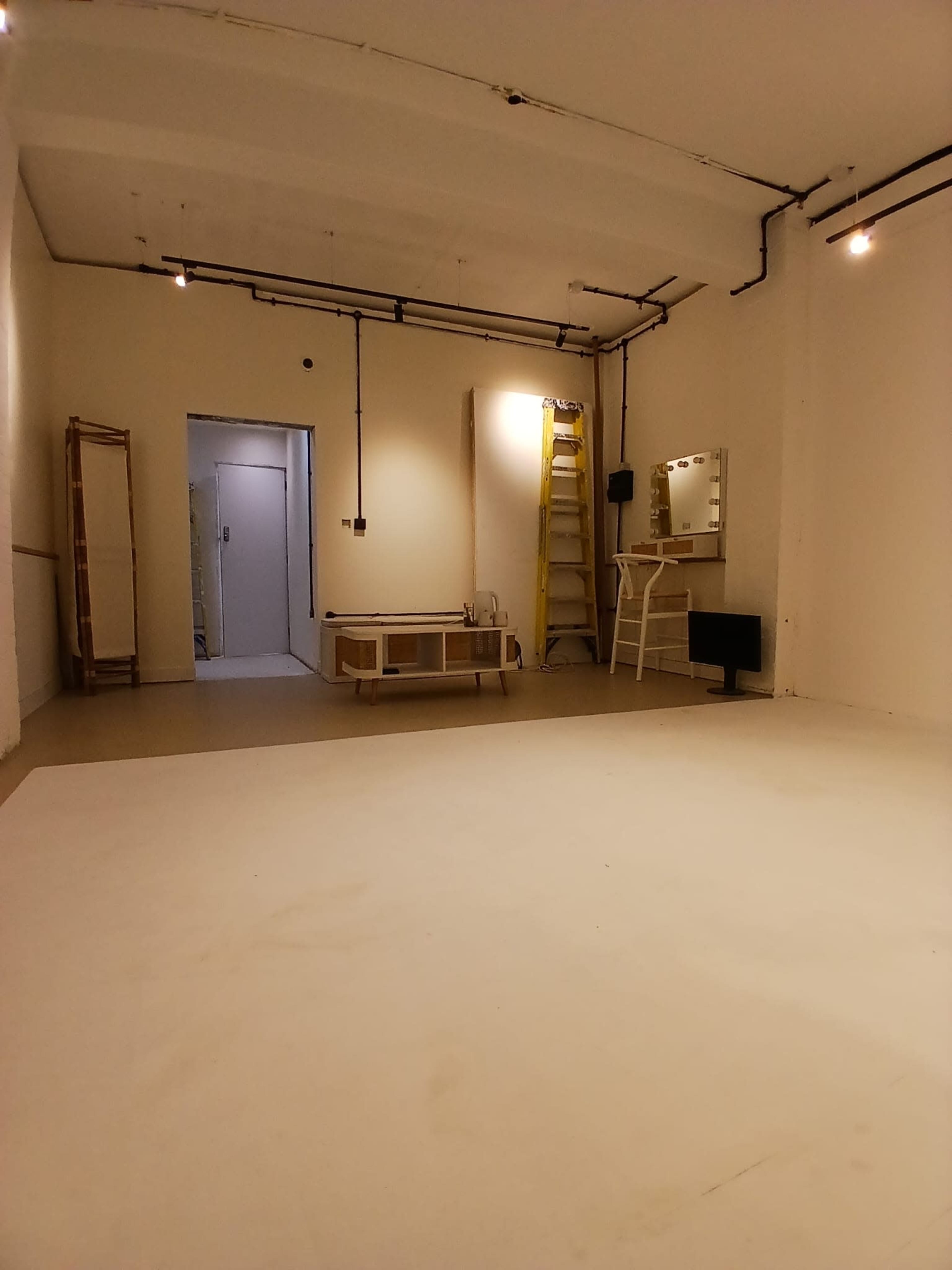 The image shows an empty room with white walls, a bare floor, a ladder against the wall, a mirror, and some furniture pieces arranged in the space.