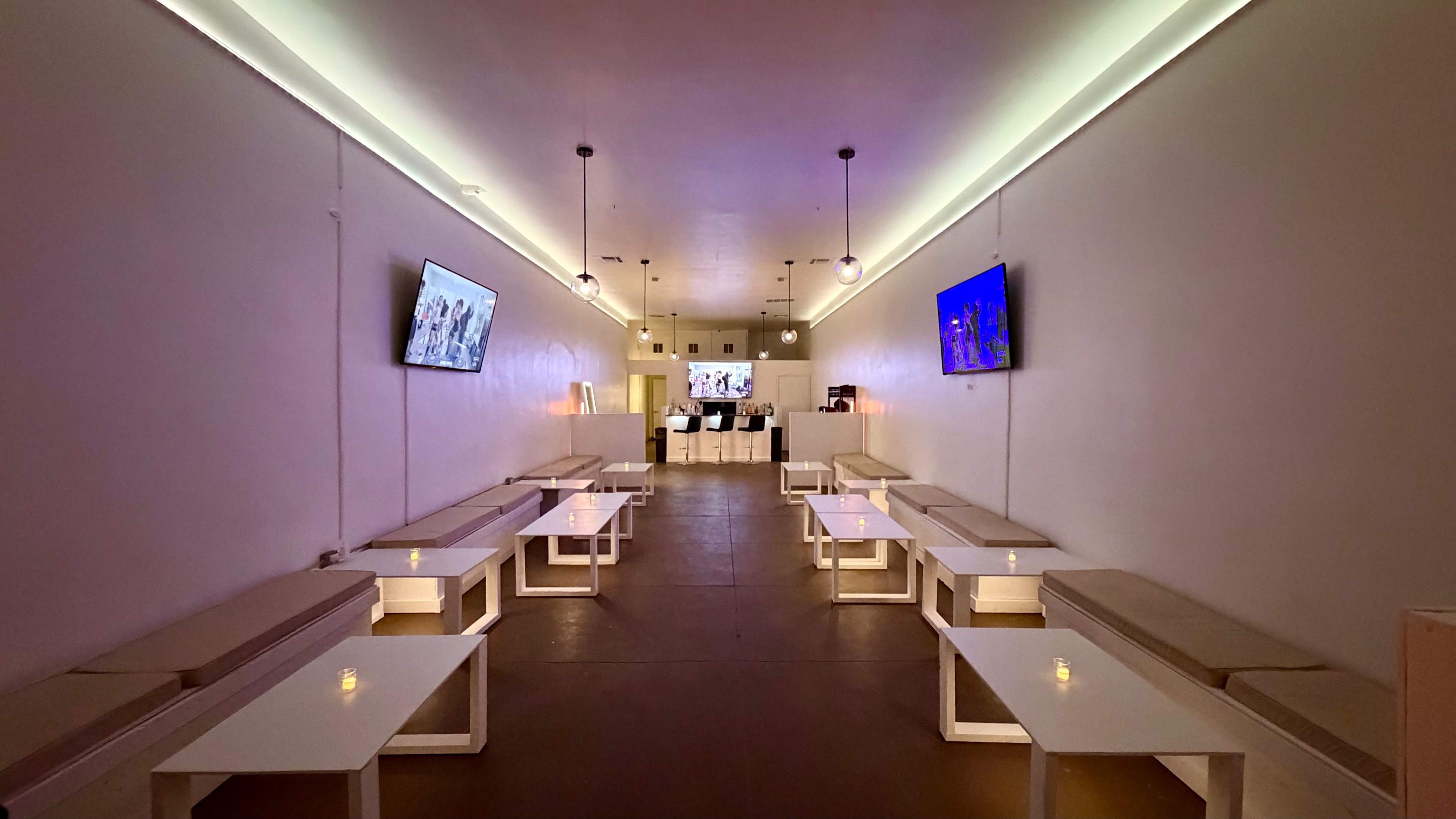 The image shows a modern lounge area with white walls, several white tables and benches, and two televisions mounted on the walls.