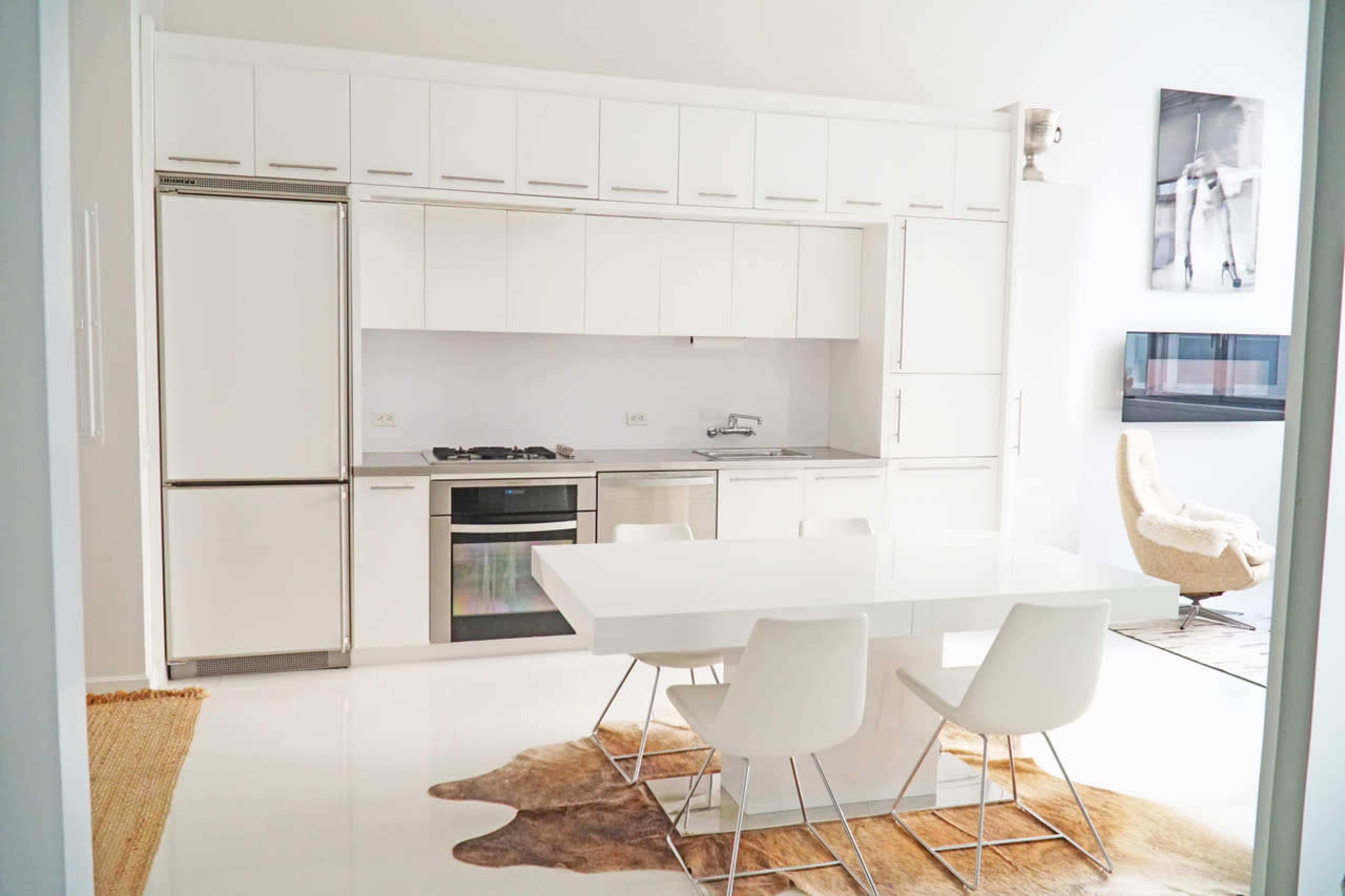 A modern kitchen with white cabinets, a stainless steel refrigerator, a stove, and a dining table surrounded by four chairs.