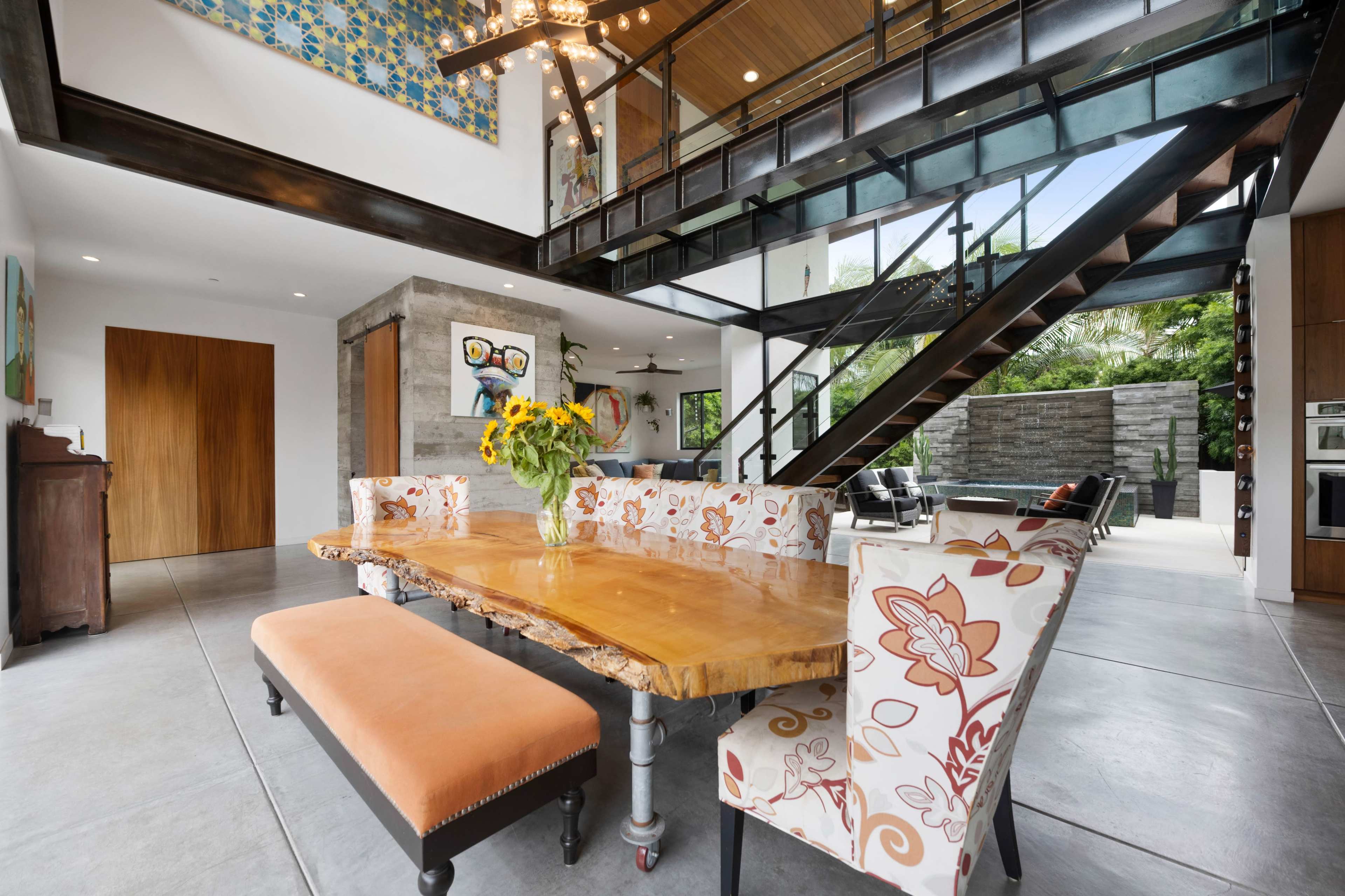 The image shows a modern dining area with a large wooden table and floral-upholstered chairs, featuring an open design that connects to an outdoor space.