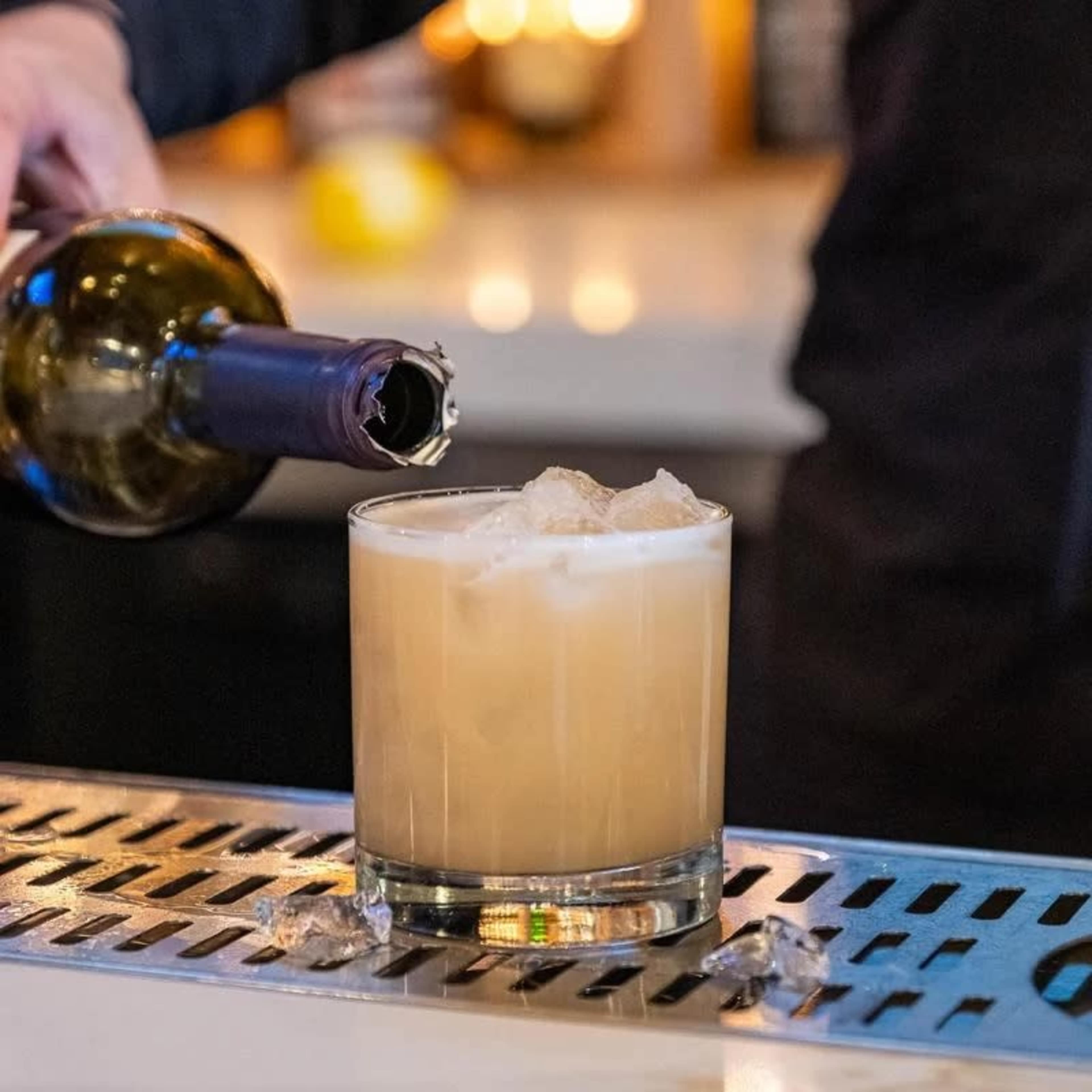A bartender pours wine into a glass filled with ice and a light-colored drink on a bar countertop.
