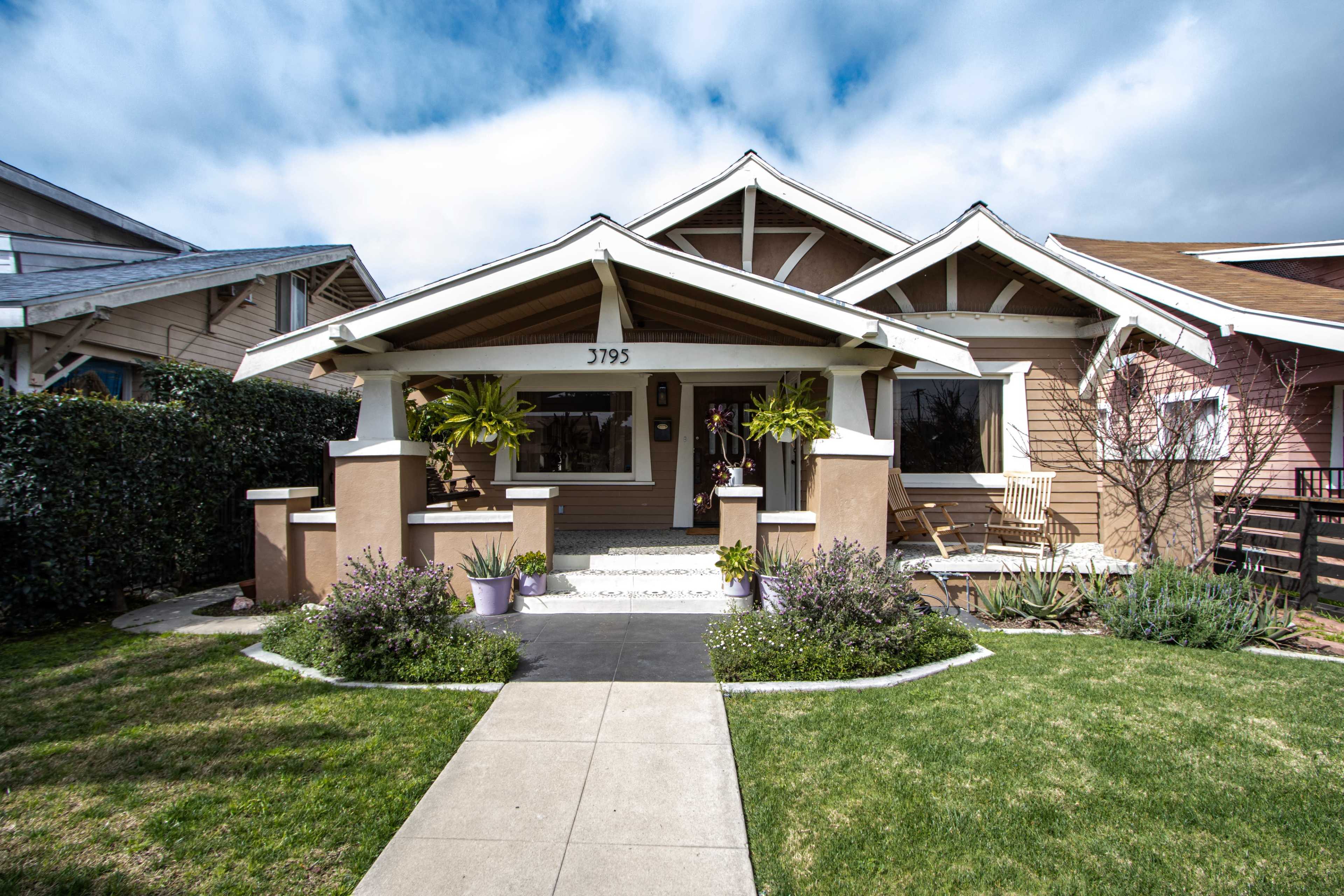 A bungalow-style house with a front porch, surrounded by landscaped greenery and a clear pathway leading to the entrance.