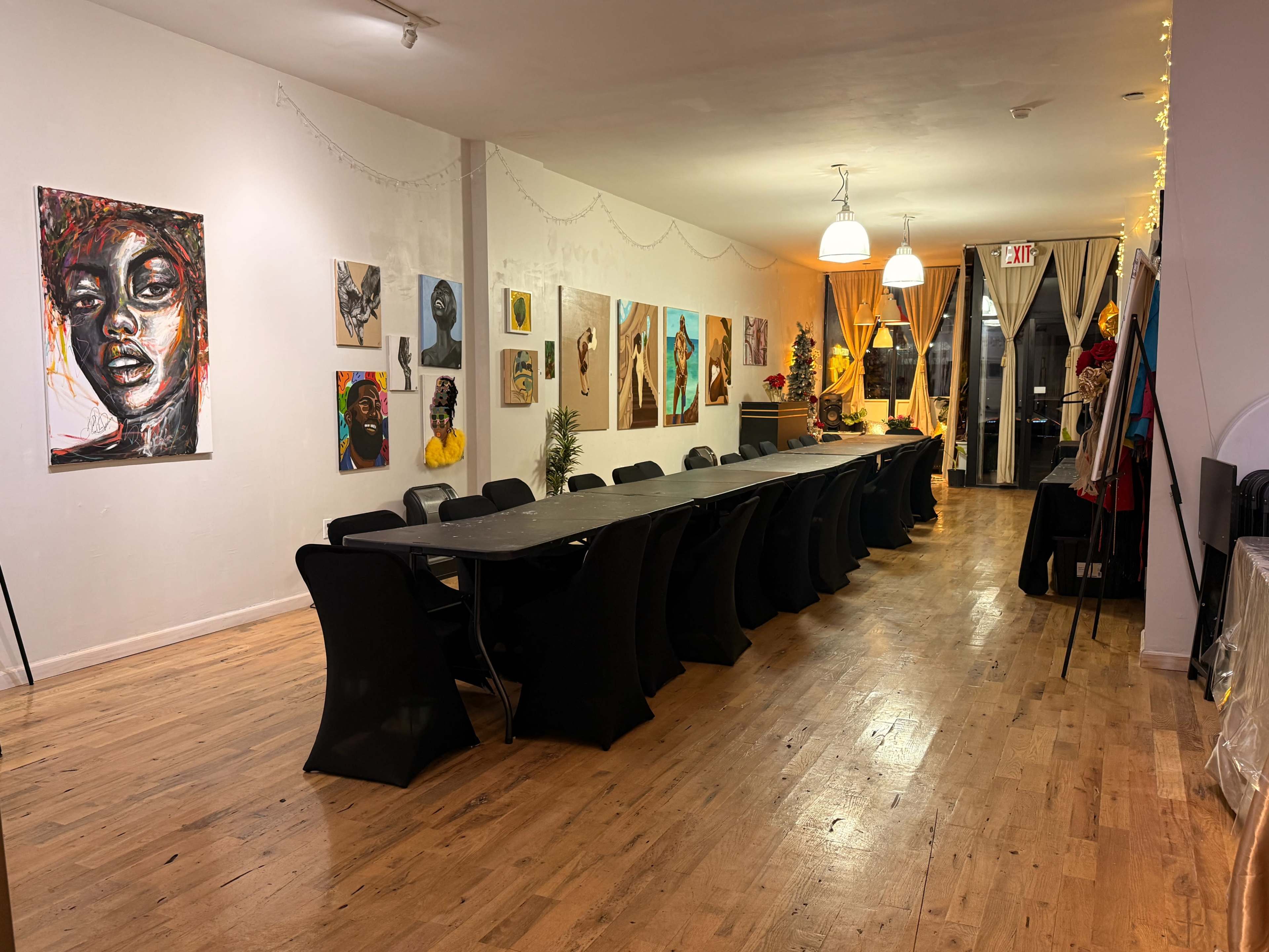 The image depicts a spacious art gallery or event space featuring a long black table surrounded by chairs, with colorful paintings displayed on the walls.
