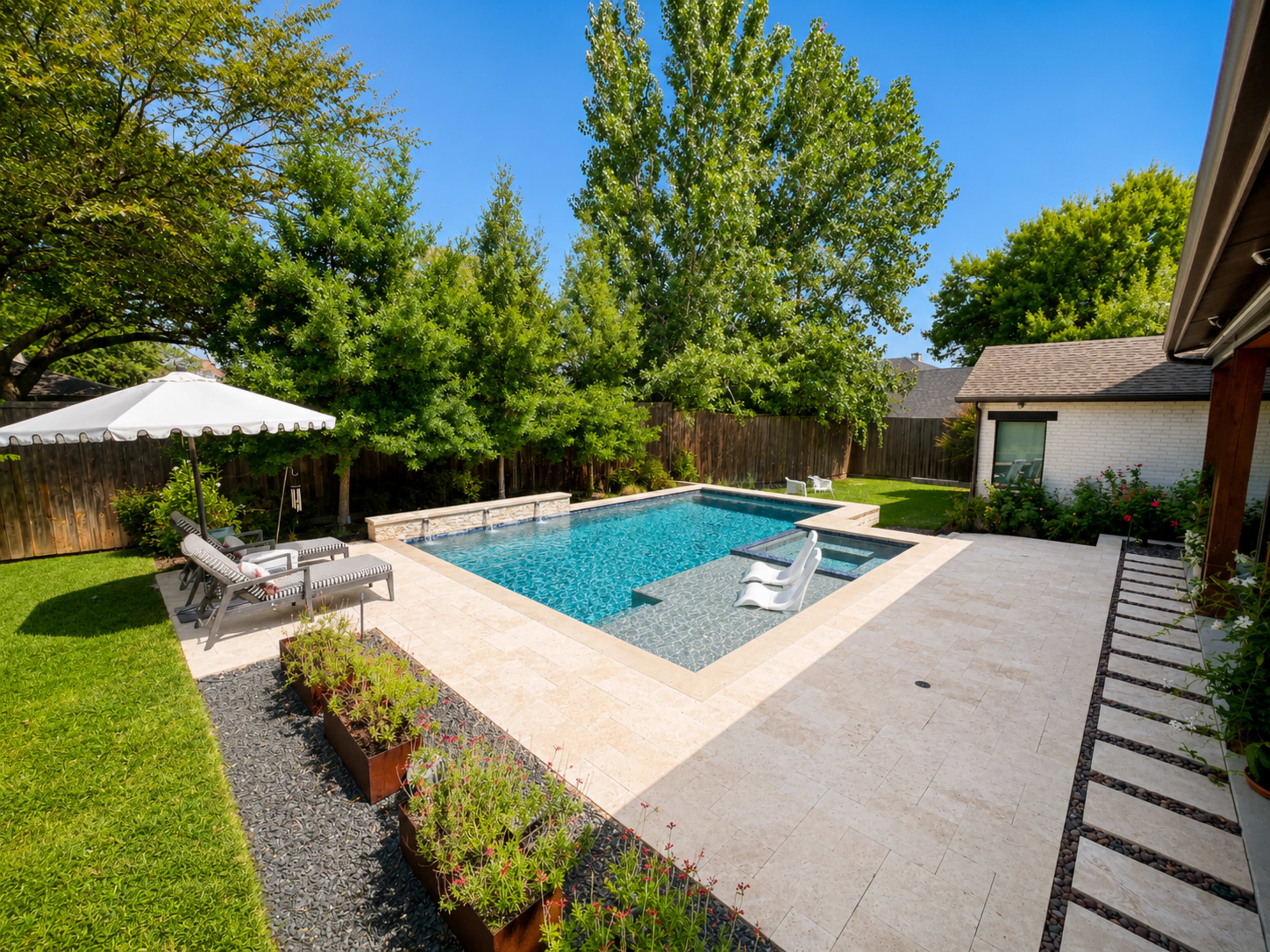 The image shows a backyard with a sparkling blue swimming pool surrounded by neatly trimmed grass, a patio area with lounge chairs, and green trees in the background.