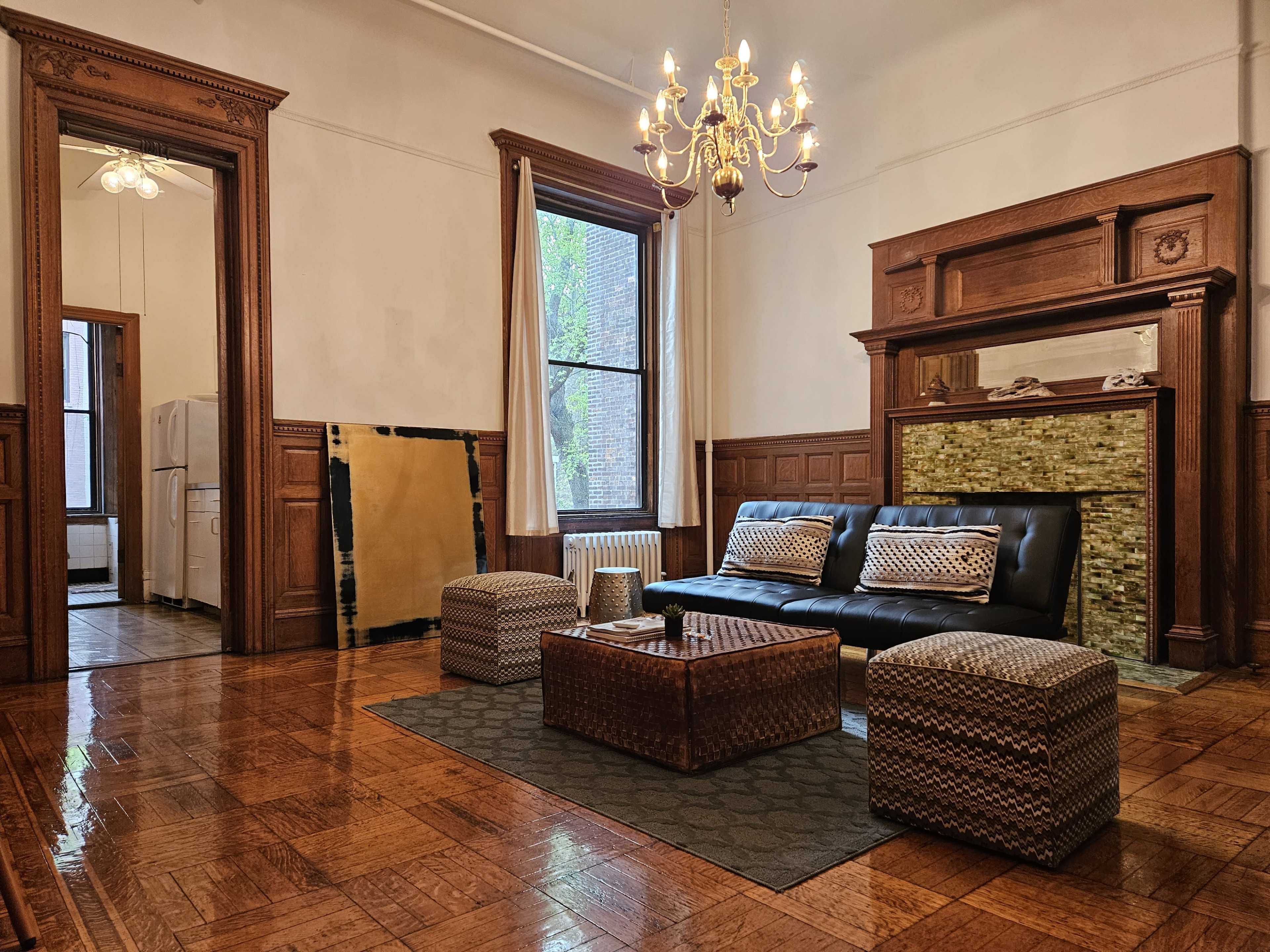 A spacious living room with a black sofa, decorative poufs, a chandelier, and a wooden fireplace surrounded by tiled accents.