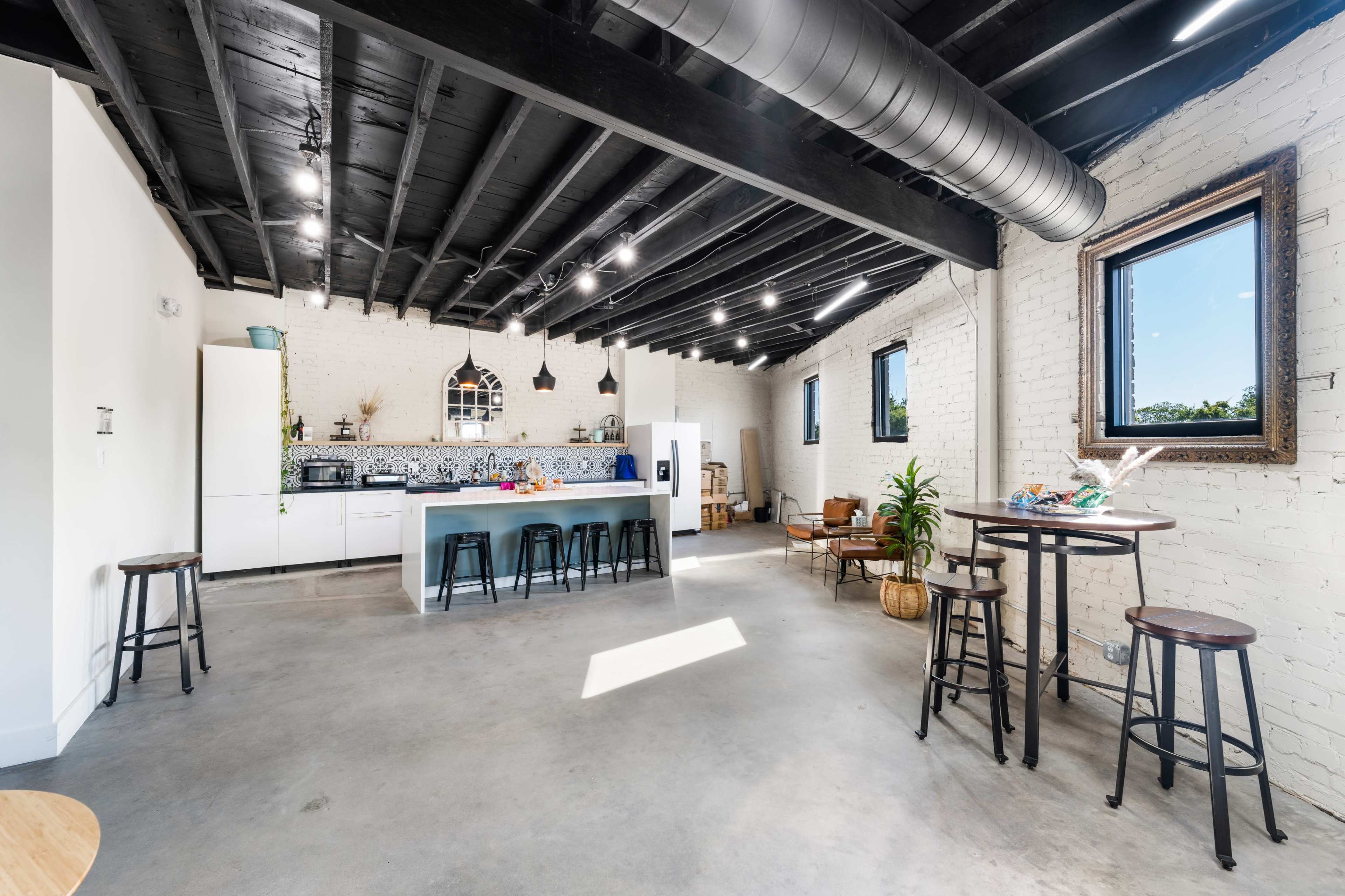 The image shows a spacious, modern kitchen and dining area with a concrete floor, exposed black beams, and large windows.