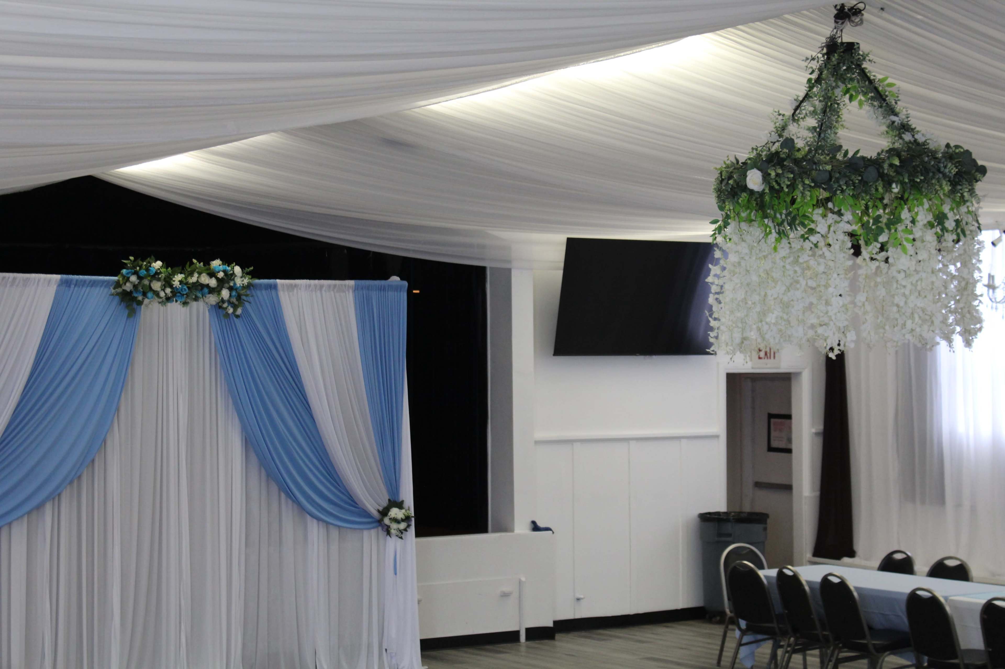 The image shows a decorated banquet hall featuring draped fabric in blue and white, floral arrangements, and seating arranged at tables.
