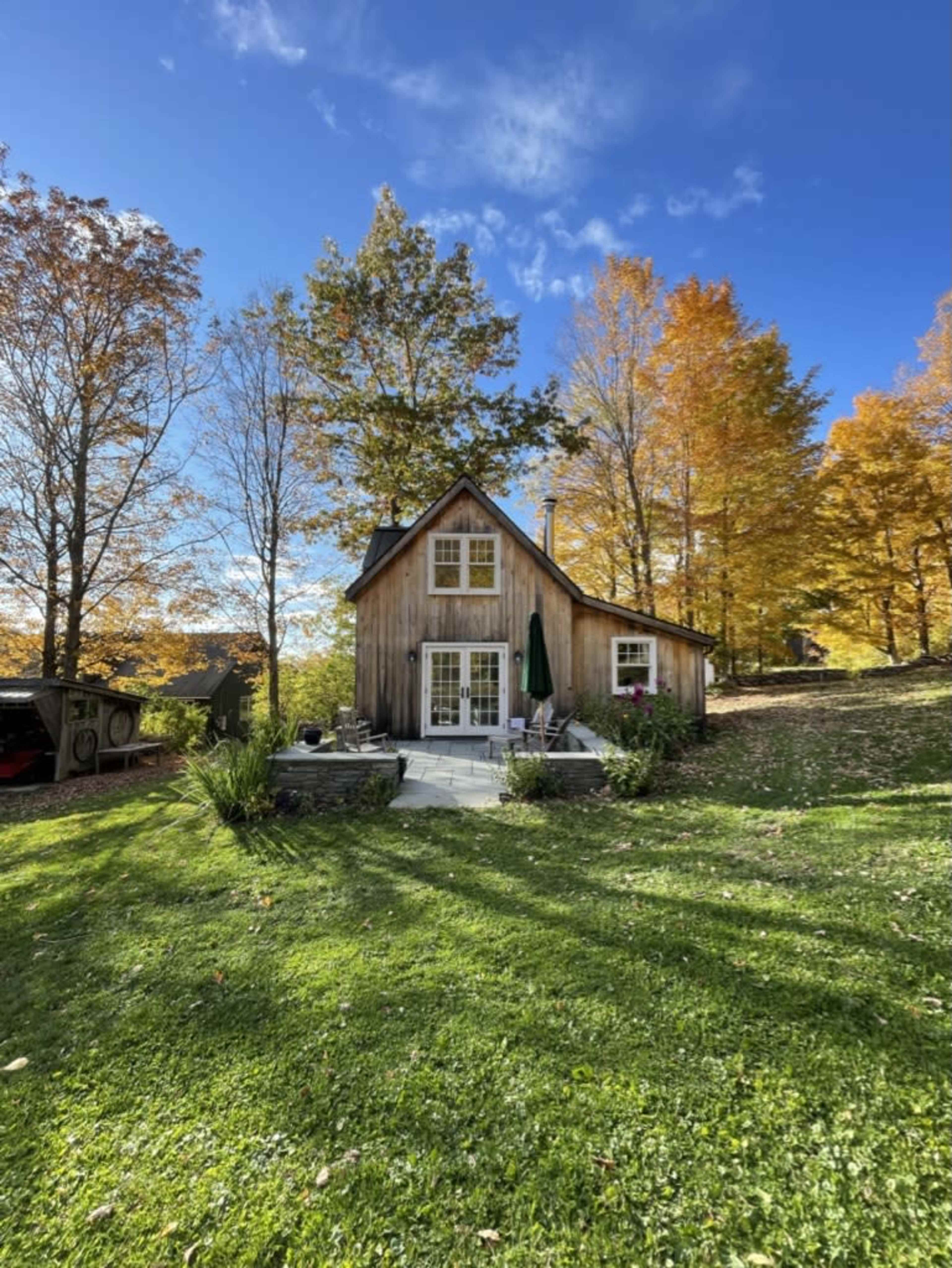 A small wooden house with a peaked roof is surrounded by colorful autumn foliage and a grassy yard.