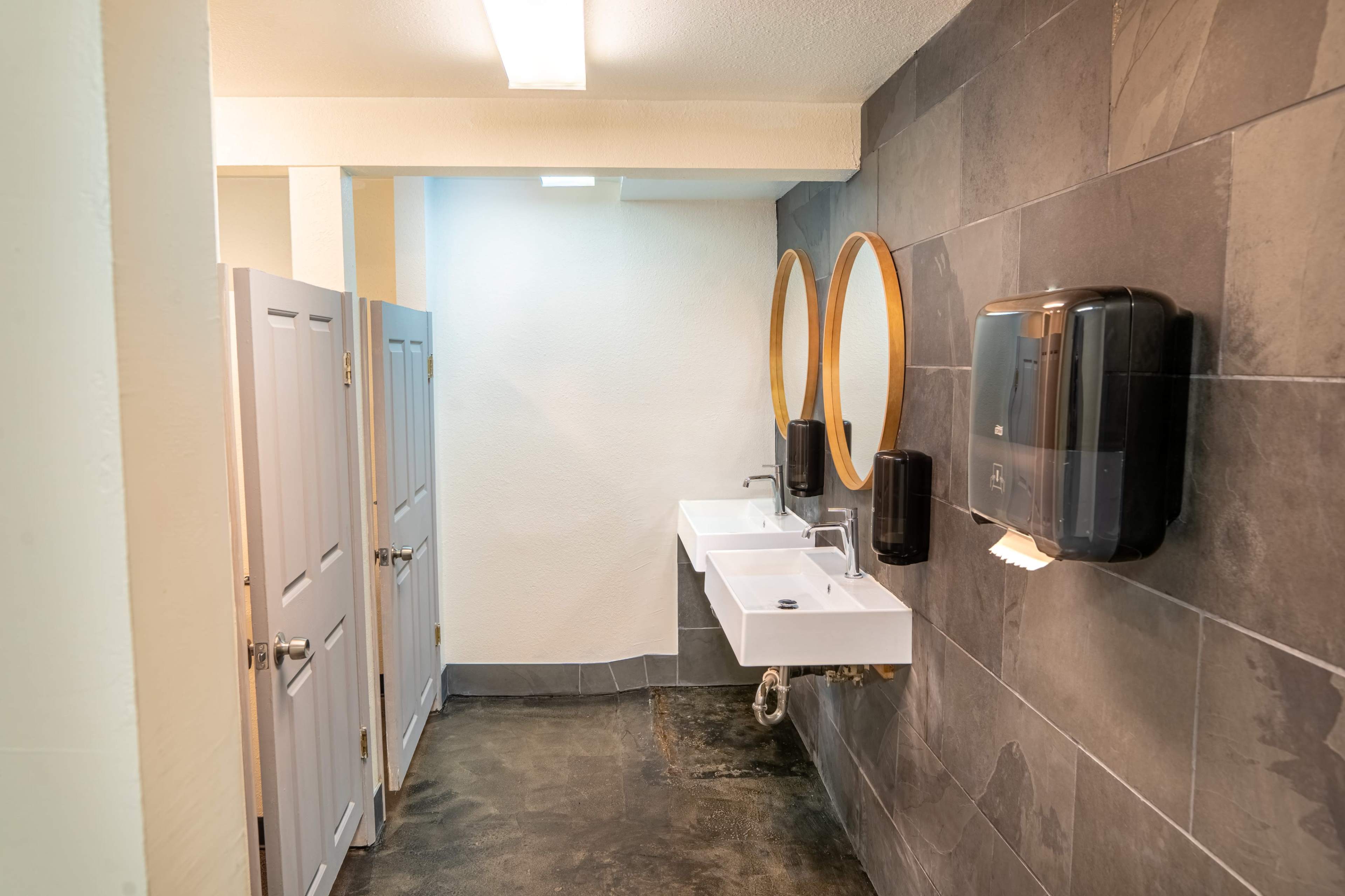 A clean bathroom with two round mirrors above a white sink and a wall-mounted soap dispenser, alongside individual stalls with white doors.