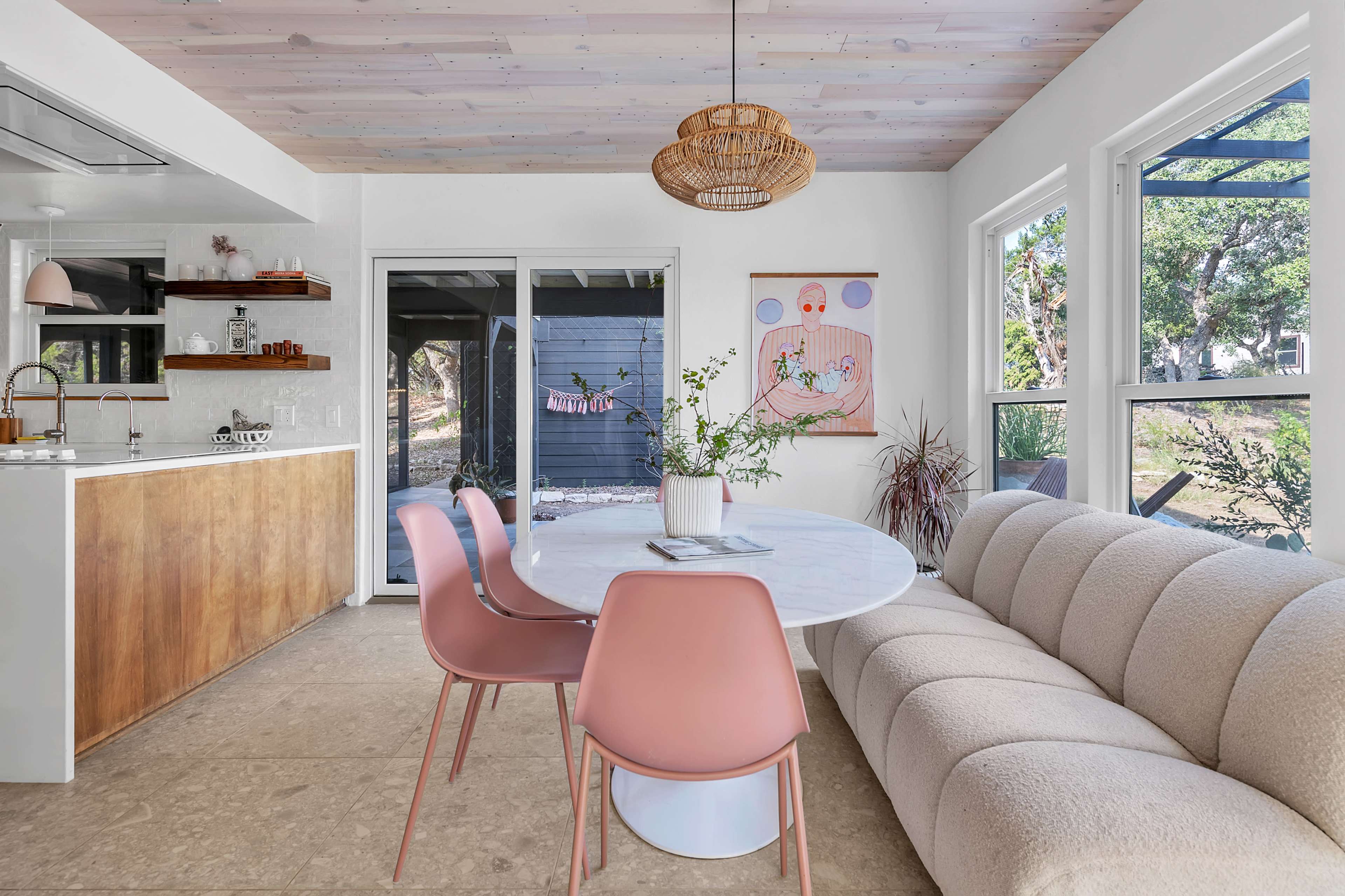 A modern dining area features a round marble table surrounded by pink chairs, with a cozy sofa and large windows allowing natural light.