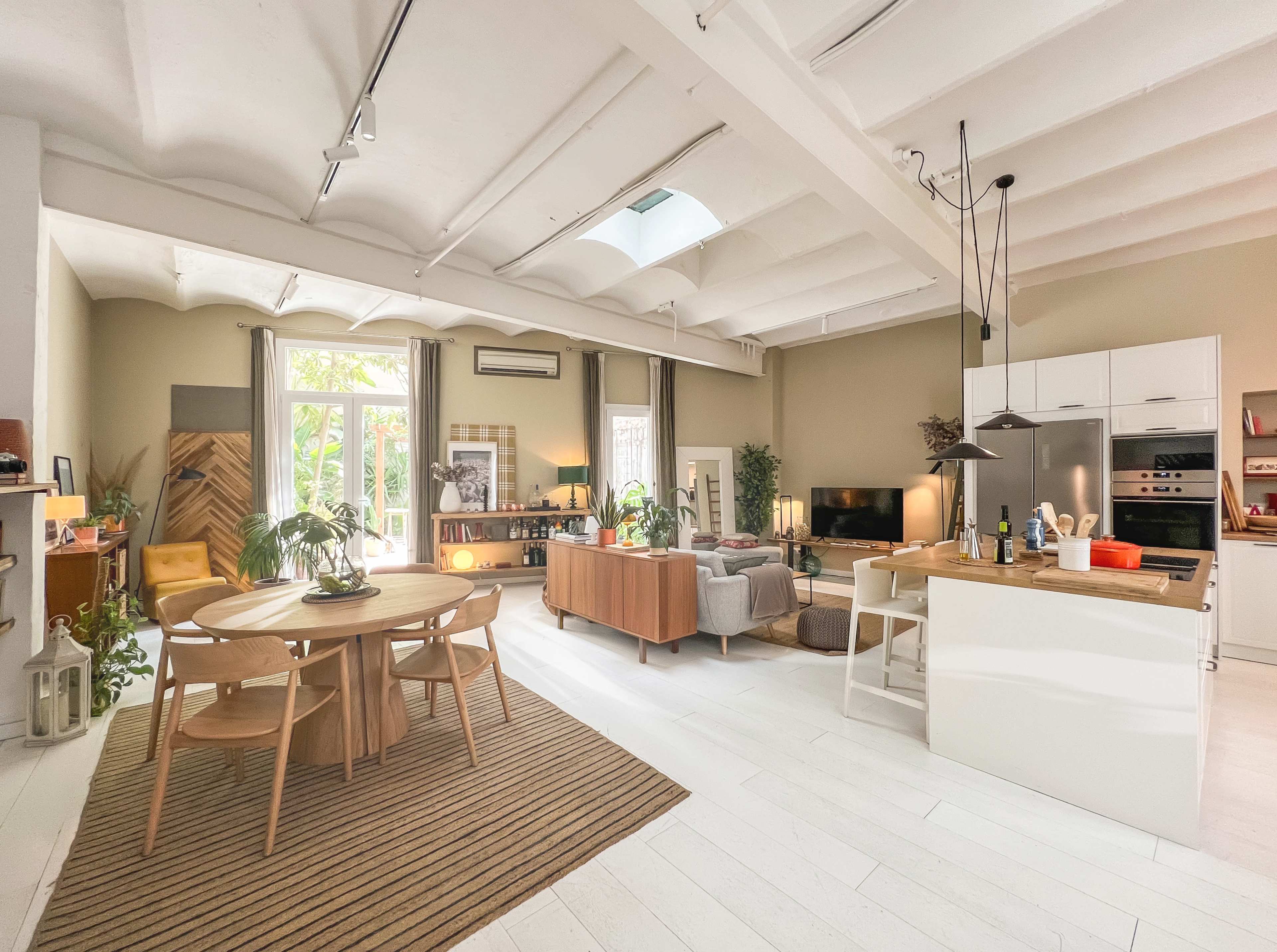 The image shows a spacious, modern living area featuring a kitchen, dining table, and seating arrangement with large windows allowing natural light to fill the room.