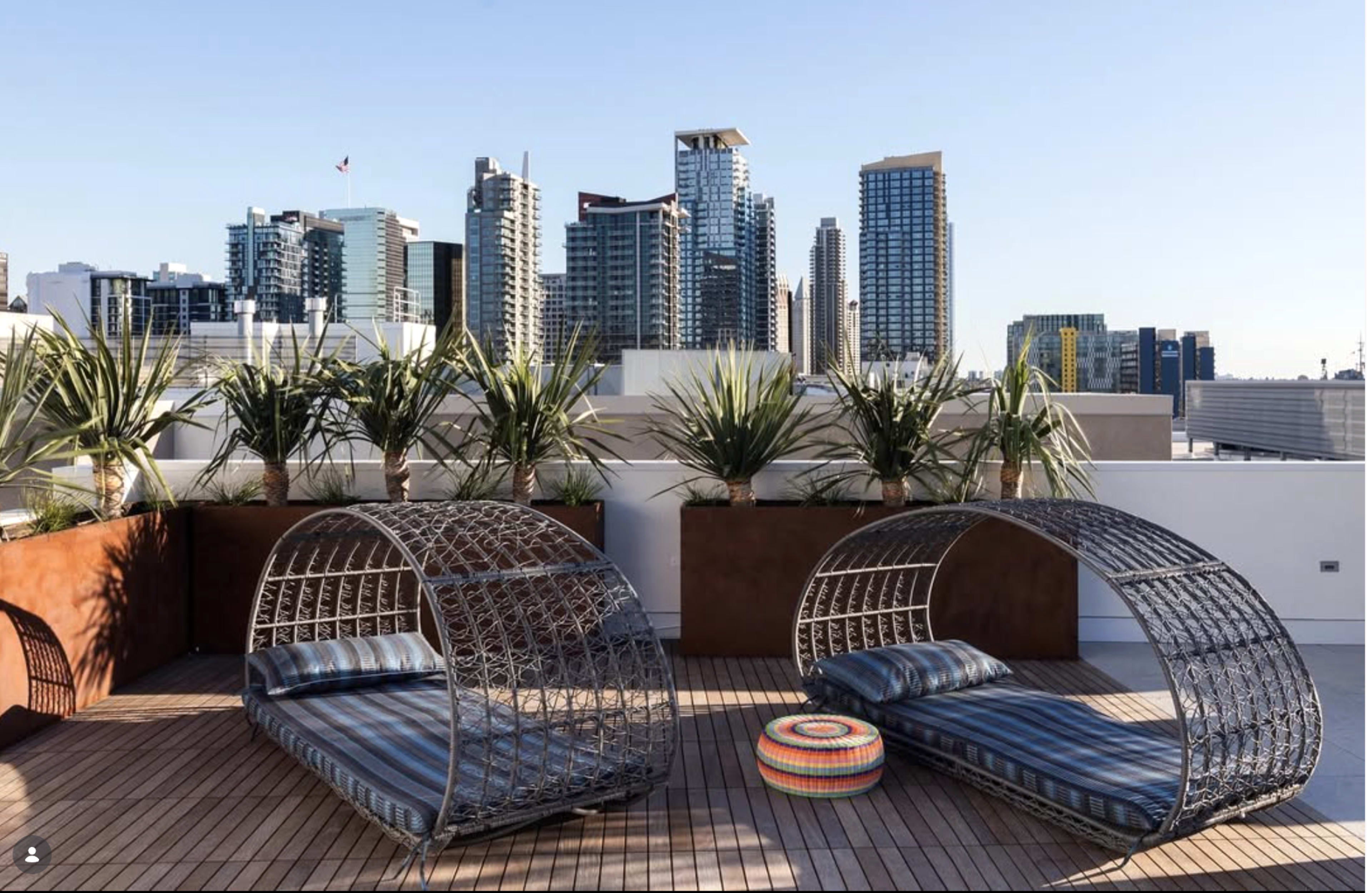 The image shows two curved lounge beds with striped cushions on a rooftop terrace, surrounded by potted palm trees and a skyline of modern buildings in the background.