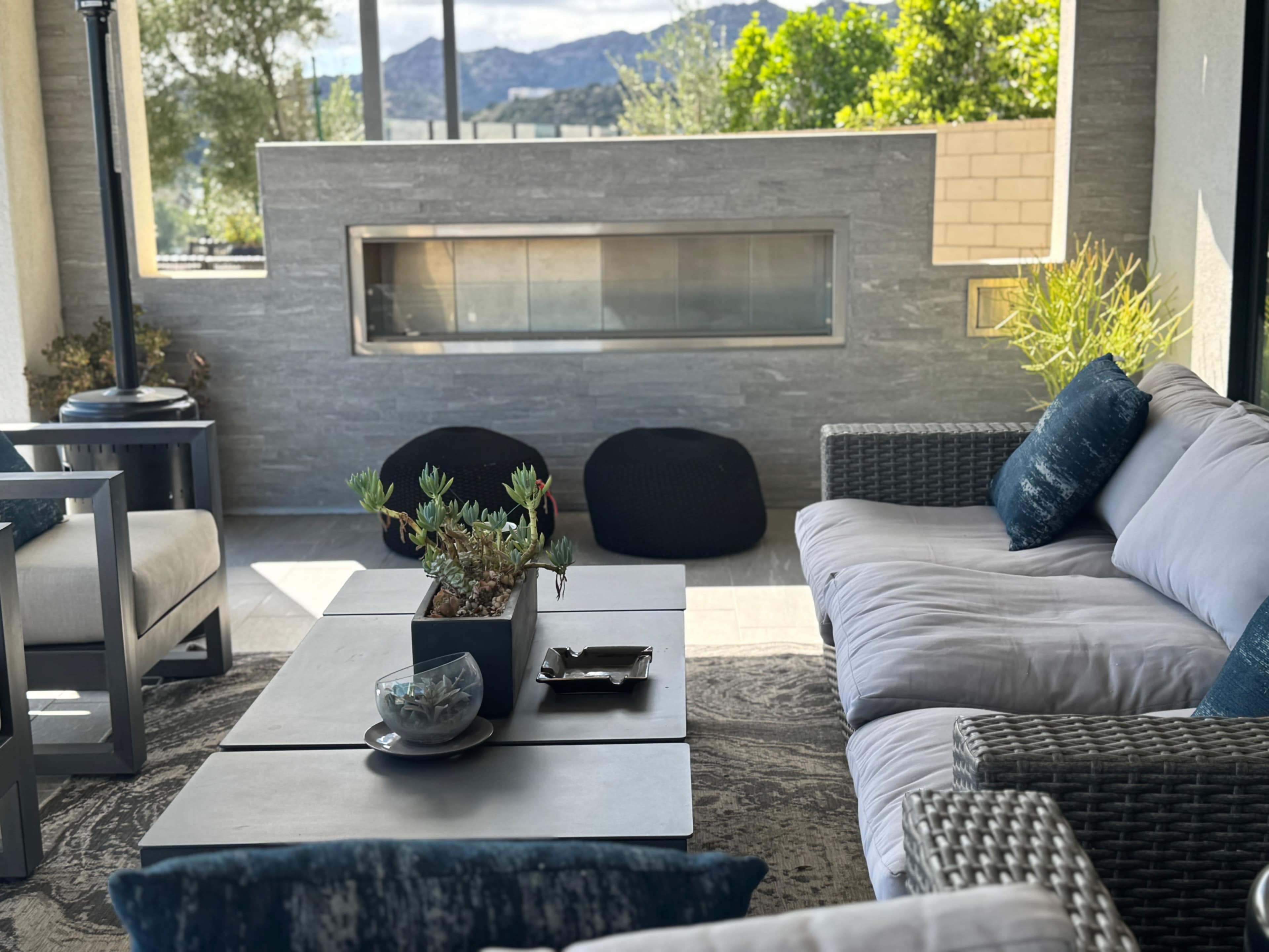 The image shows a modern outdoor living space featuring a gray stone wall with a fireplace, a table with decorative items, and comfortable seating arranged around the area.