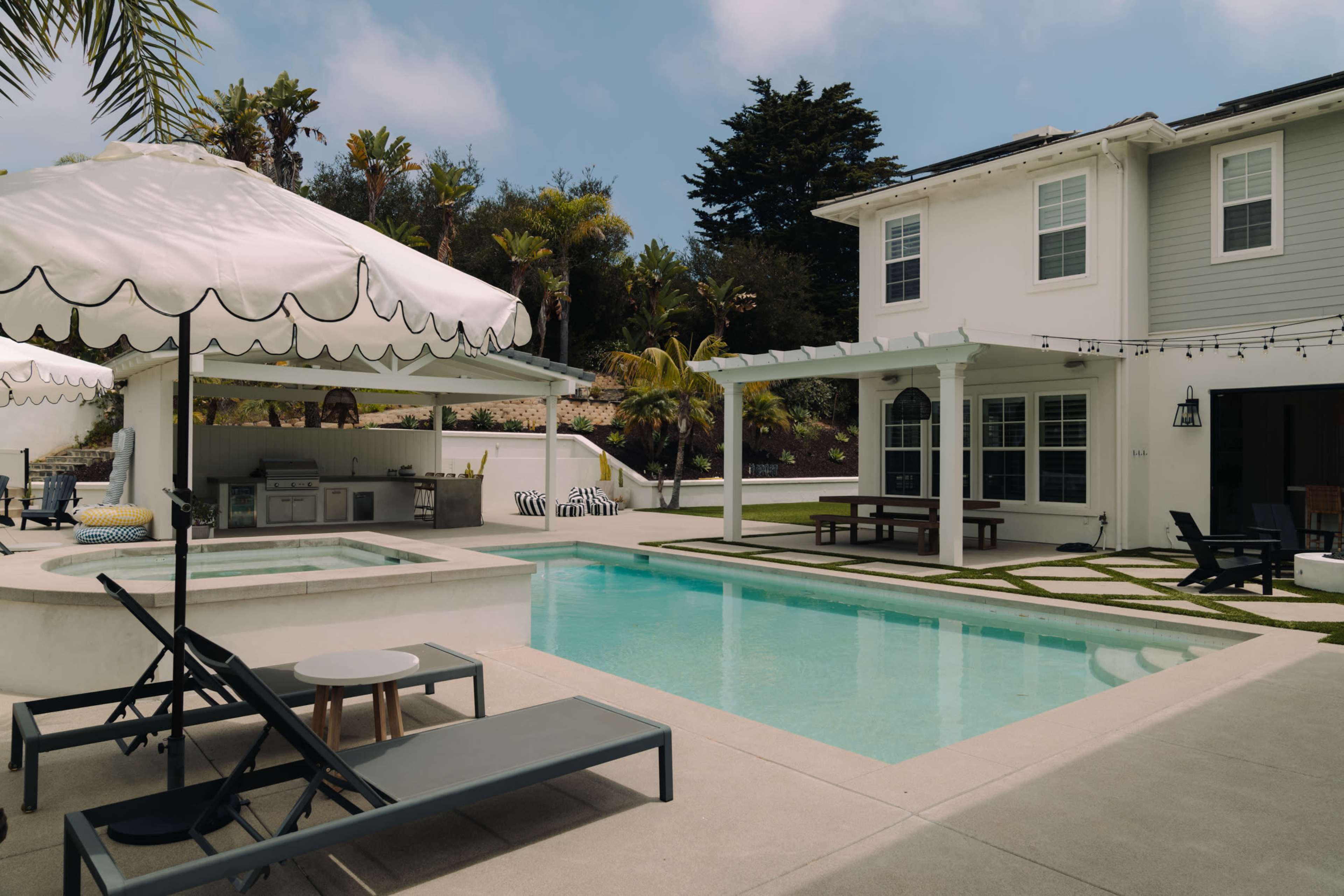 The image shows a modern backyard with a swimming pool, lounge chairs, an outdoor kitchen, and a large covered seating area next to a two-story house.