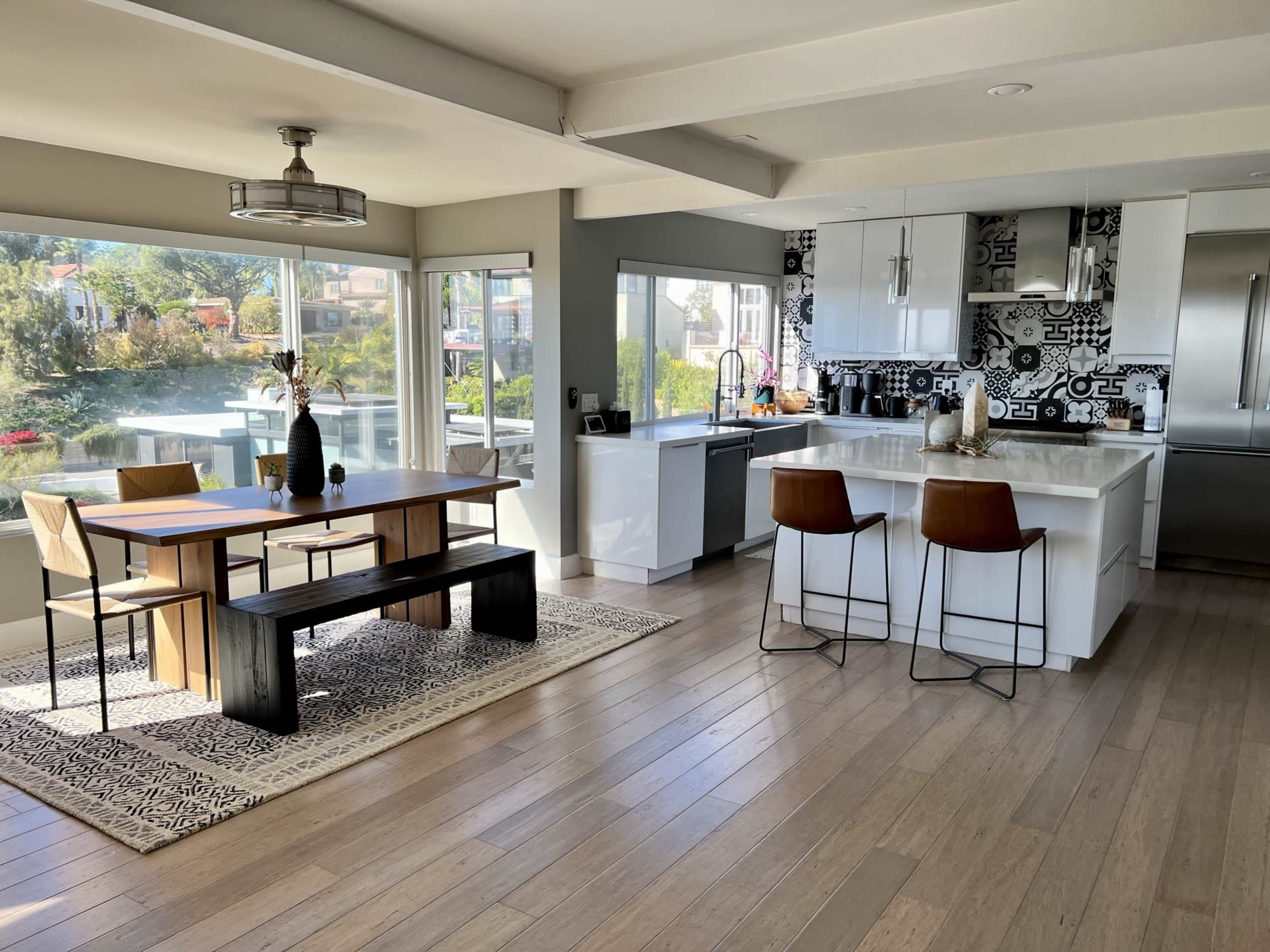 The image shows a modern kitchen and dining area with a wooden table, stools, and large windows overlooking a body of water.
