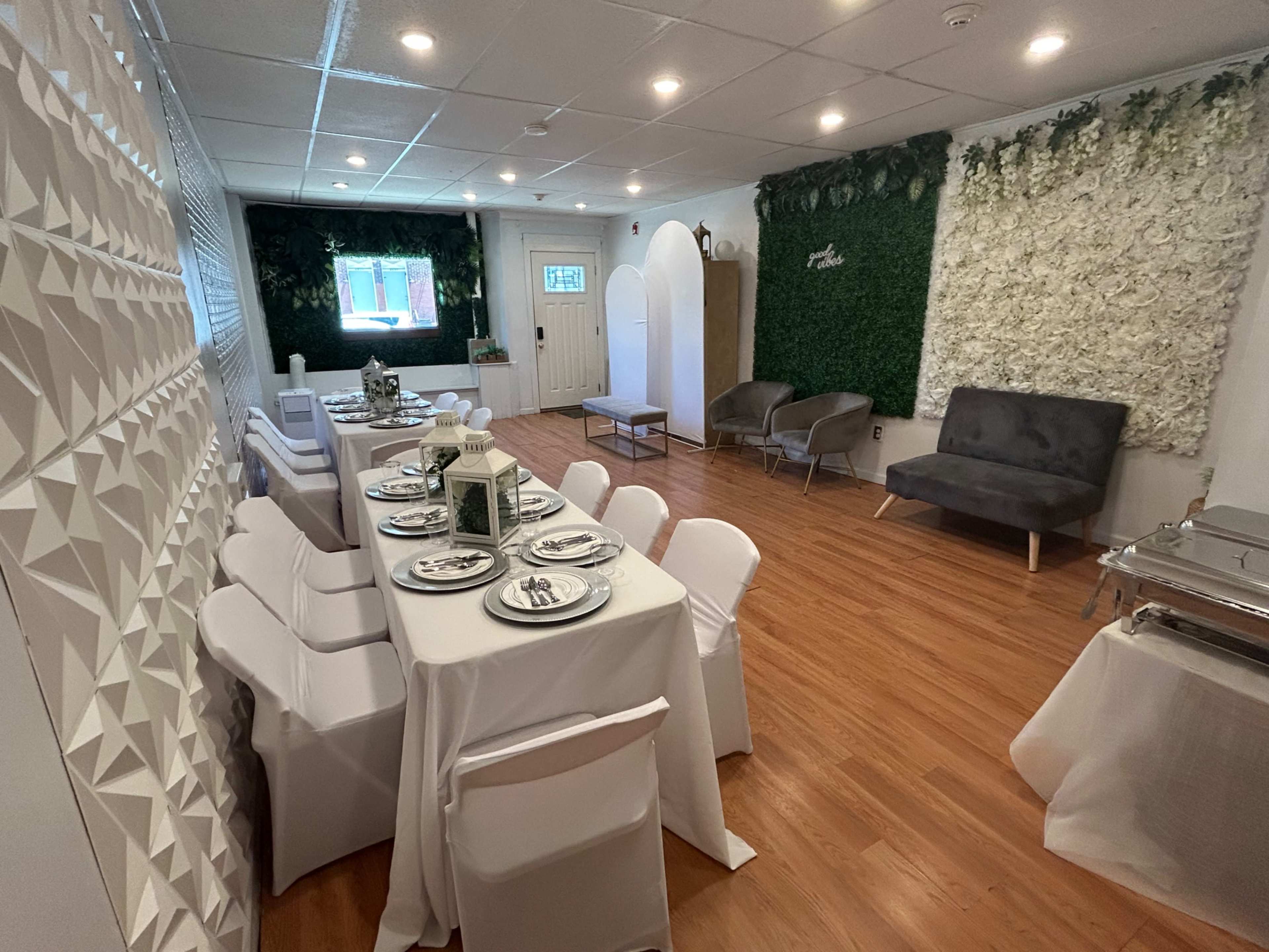 The image shows a dining area with a long table set for a meal, surrounded by white chairs, greenery on the walls, and a cozy seating area.