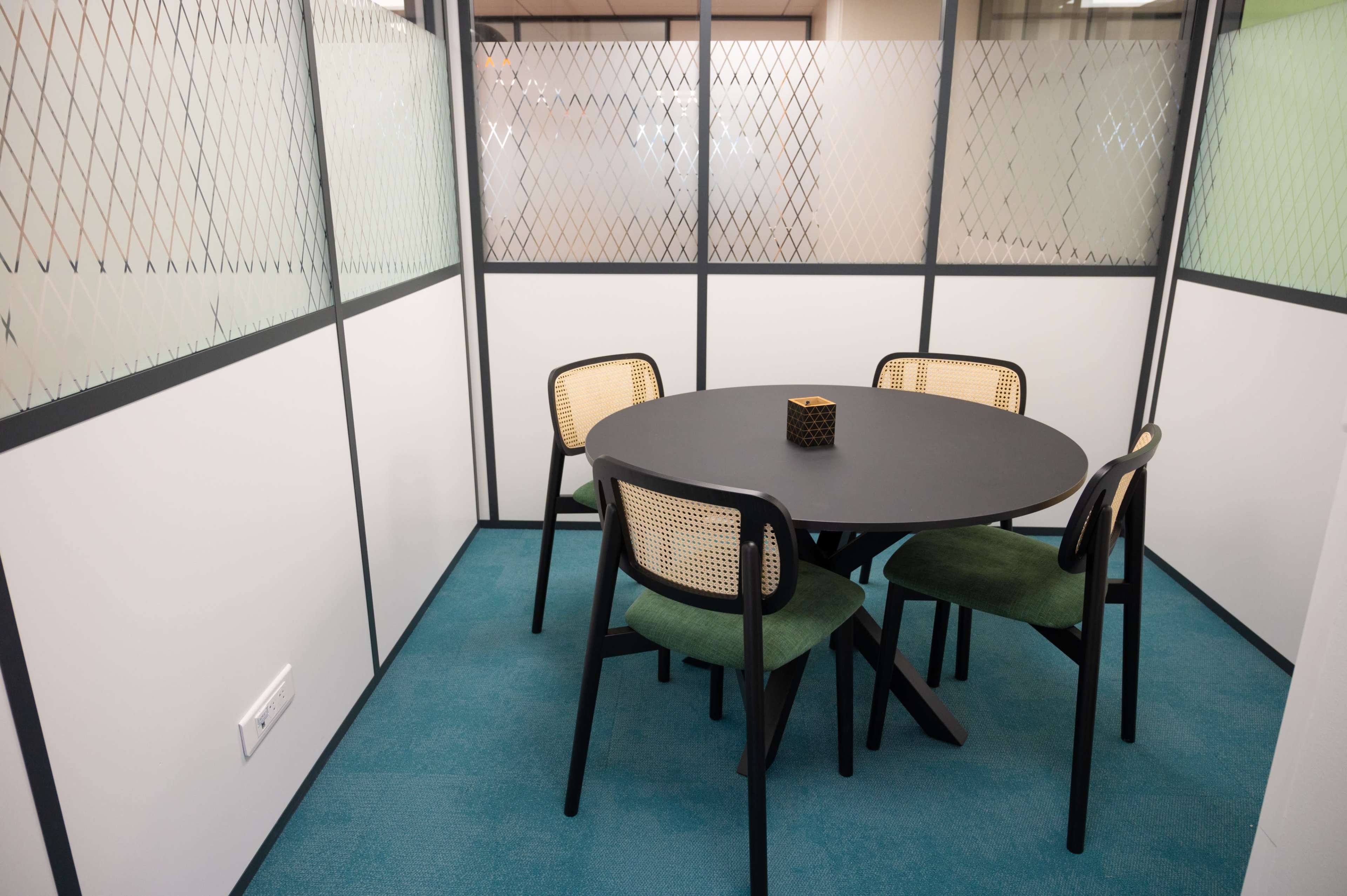 A compact meeting room features a round black table surrounded by four chairs, enclosed by glass partitions with a diamond pattern.