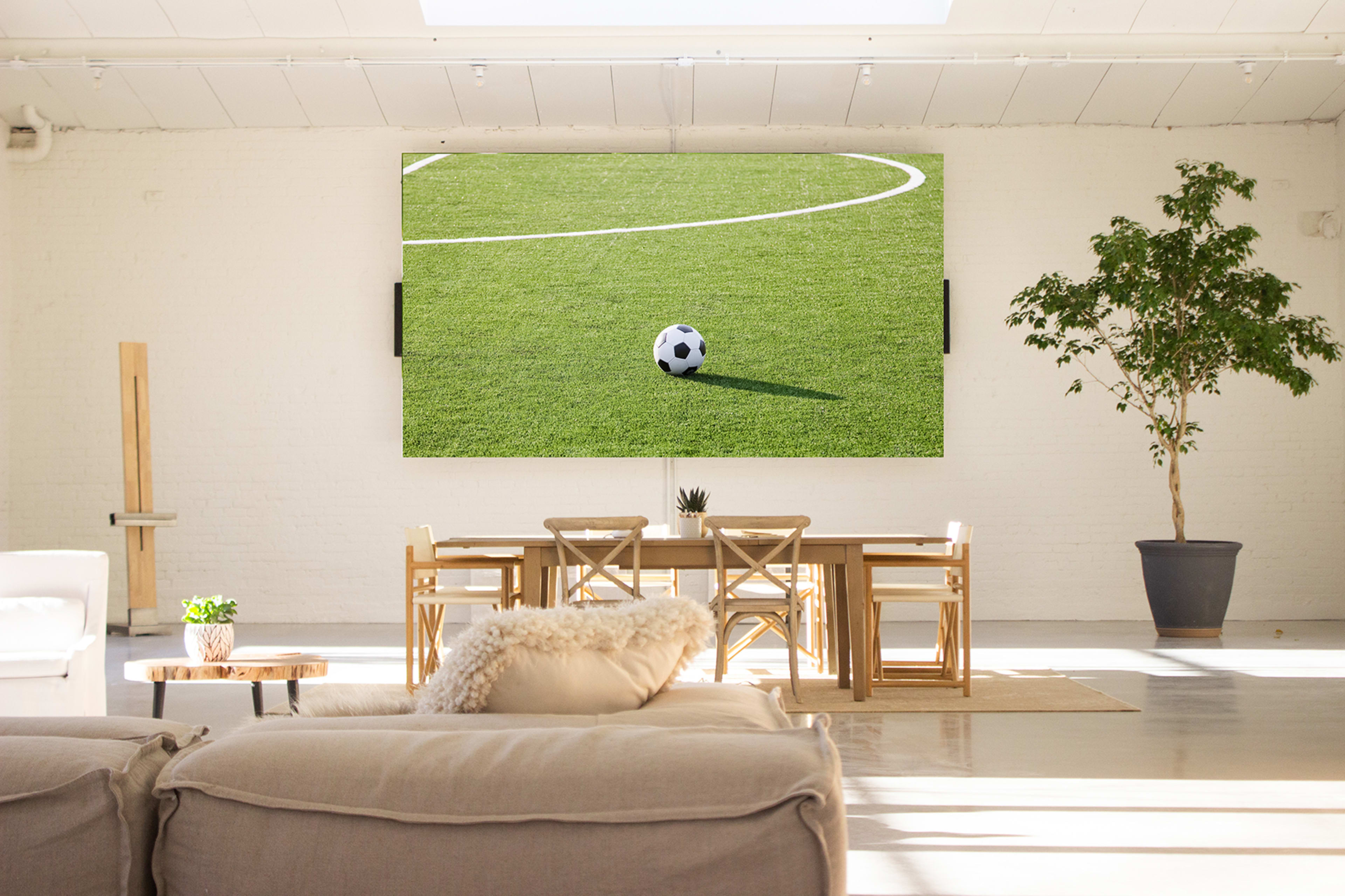 A large TV displaying a soccer ball is mounted on a wall in a bright, modern living space with a dining table and plants.