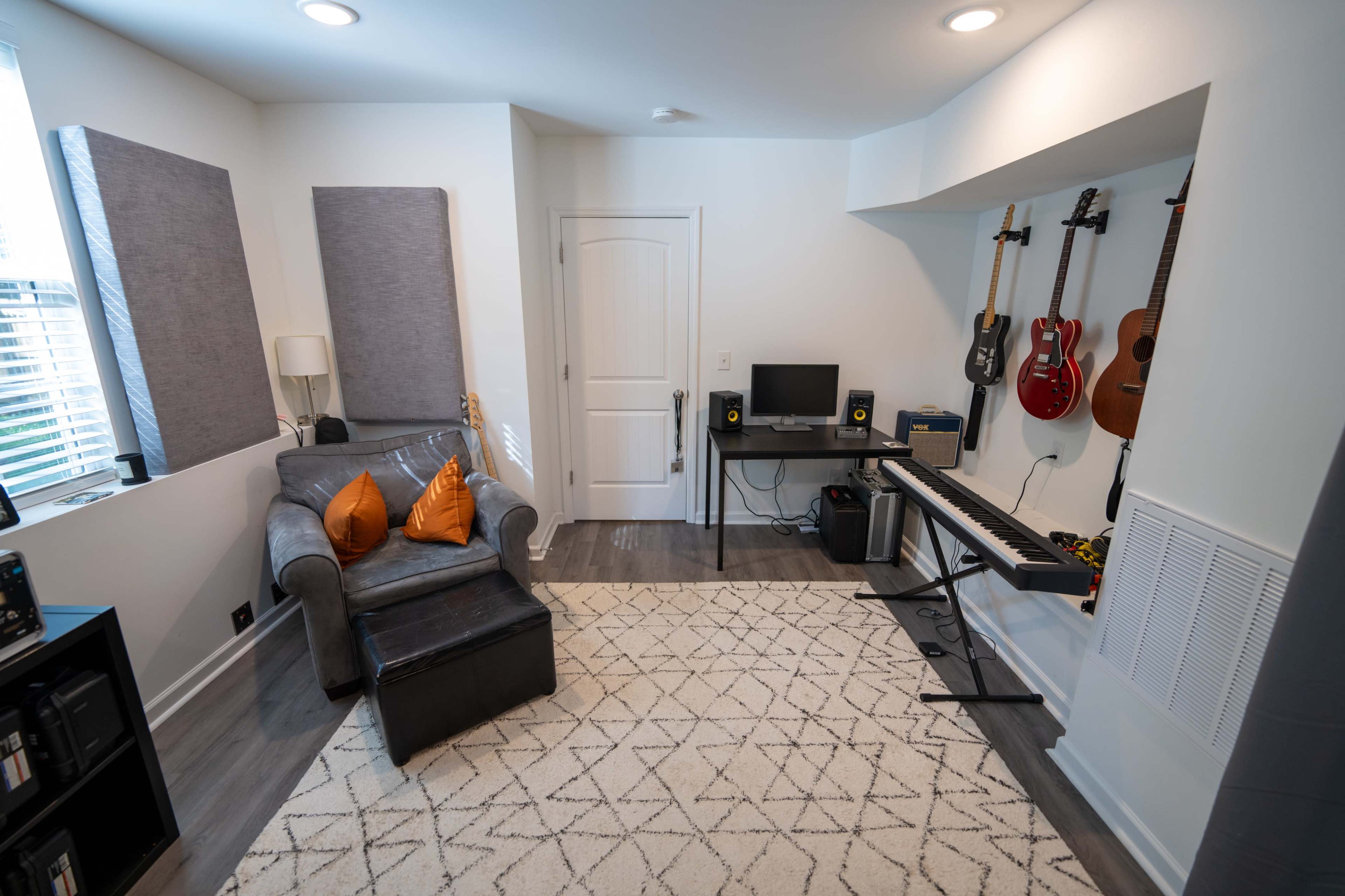 The image shows a music room with a gray couch, a black ottoman, a computer desk with speakers, a keyboard, and several guitars mounted on the wall.
