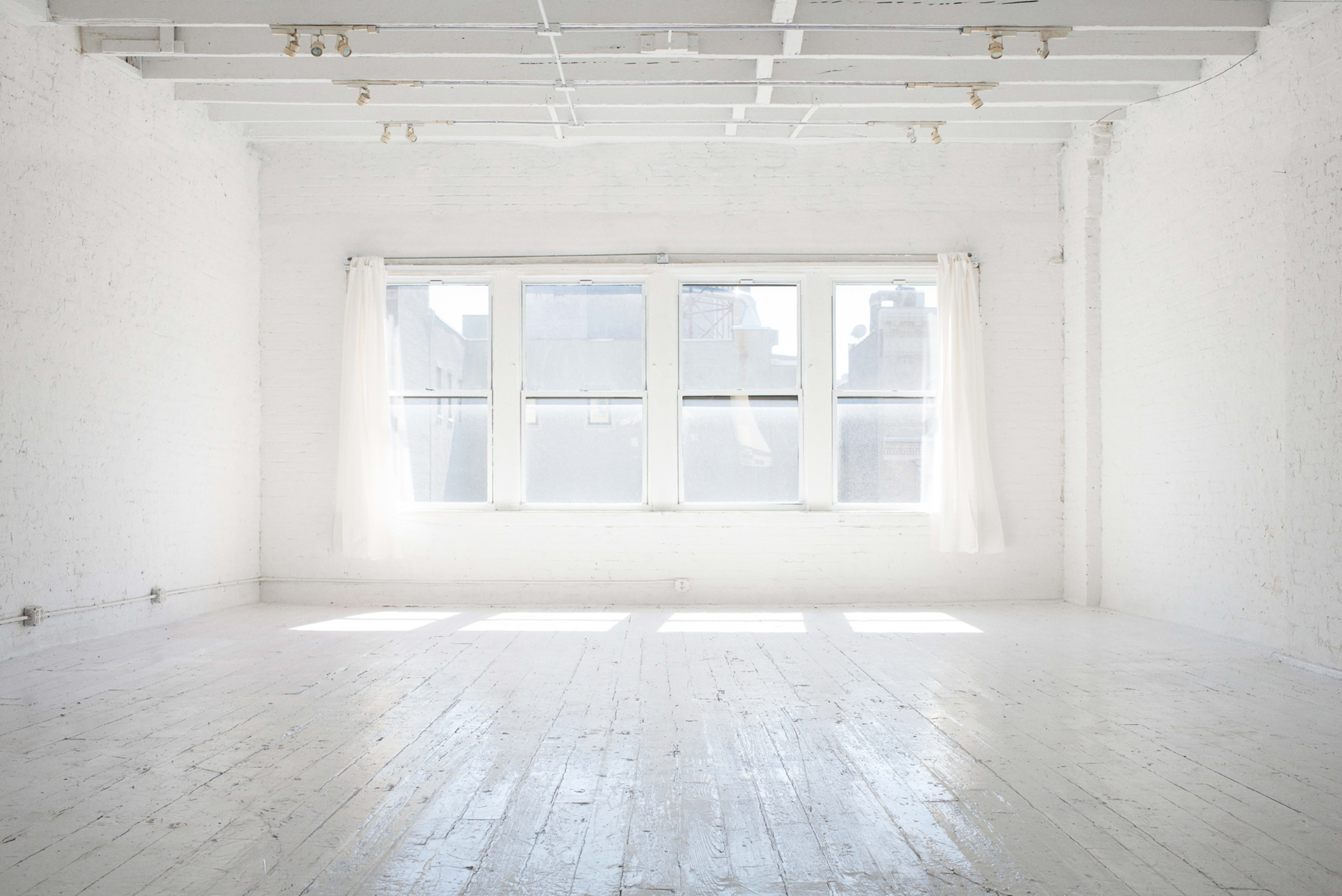 A bright, empty room with white walls and wooden flooring, featuring large windows with sheer curtains that allow light to fill the space.