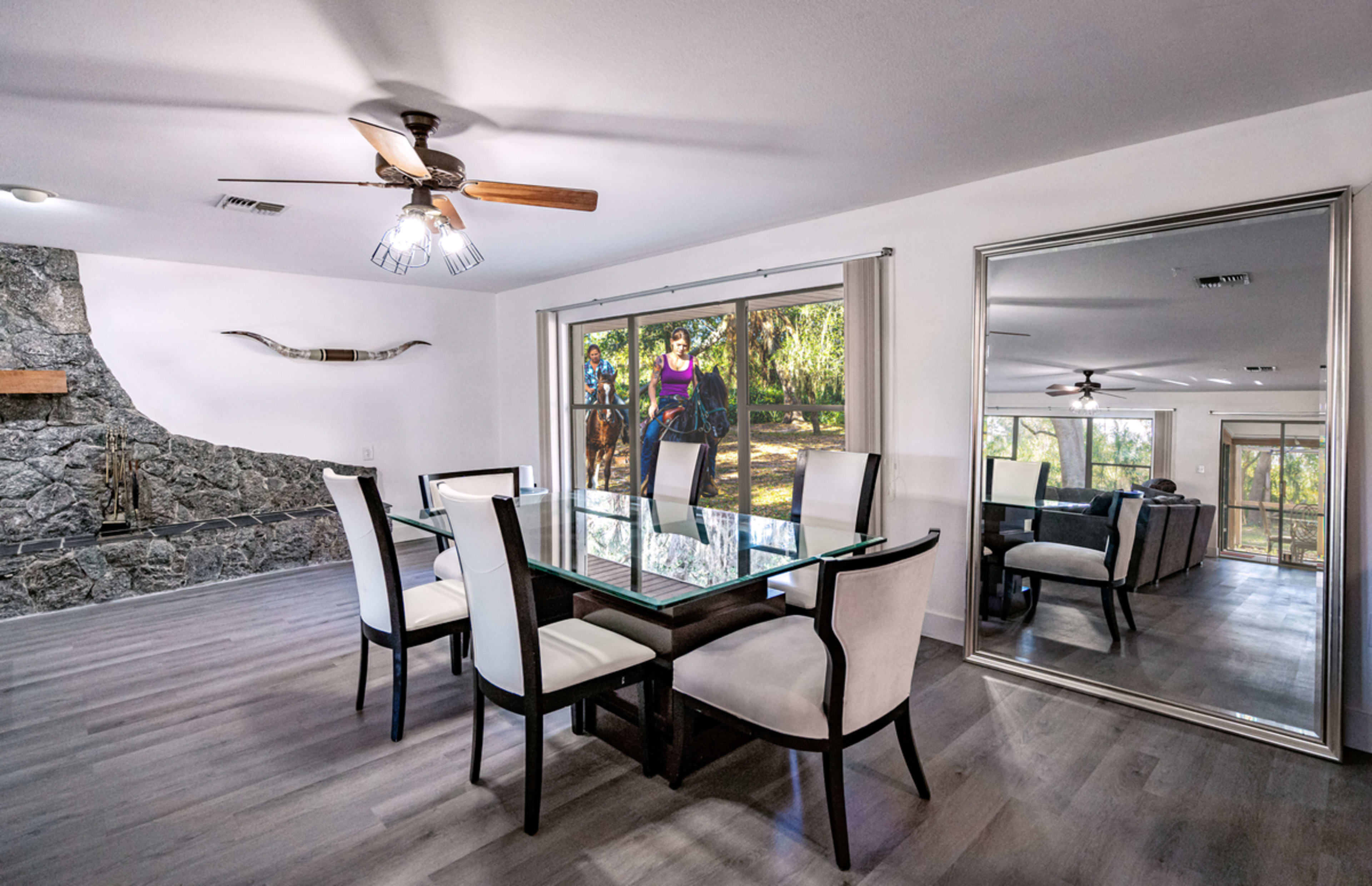 A dining area features a glass table surrounded by six chairs, with a large window showing two people on horseback outside.