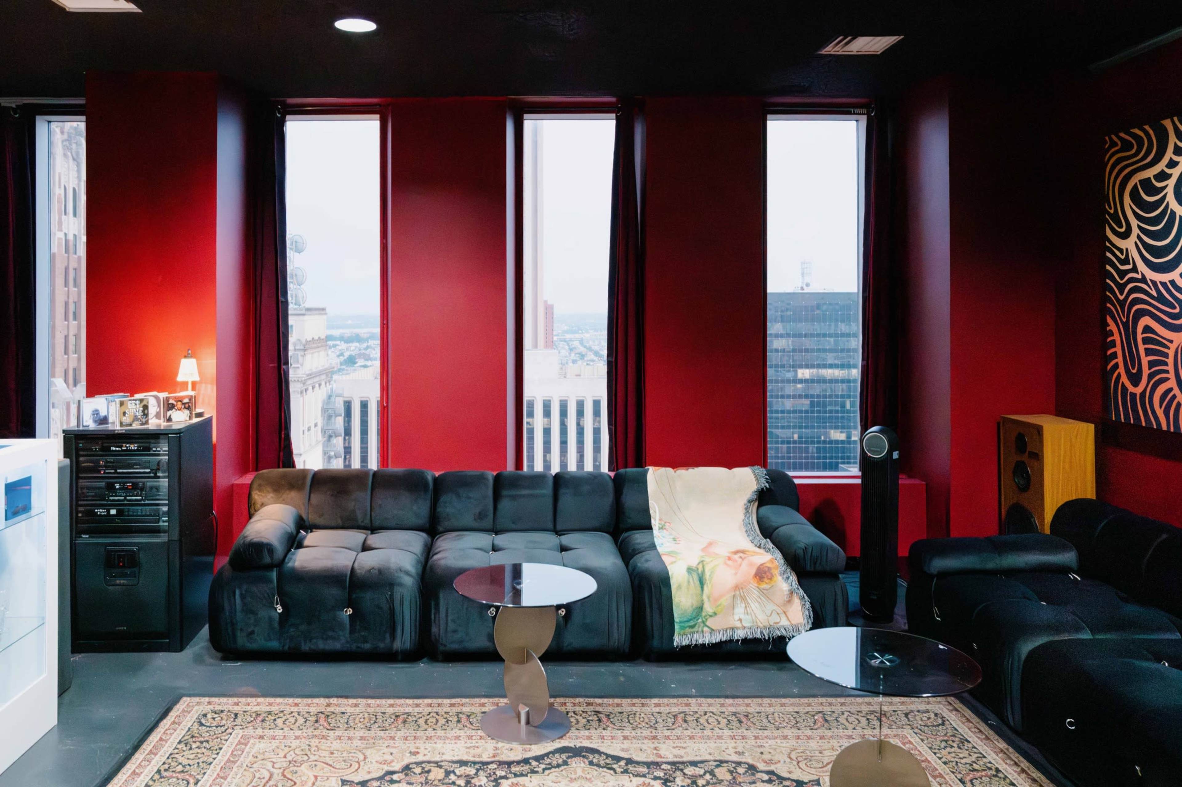The image shows a modern living space with dark red walls, large windows, a black sectional sofa, two small tables, and a decorative rug.