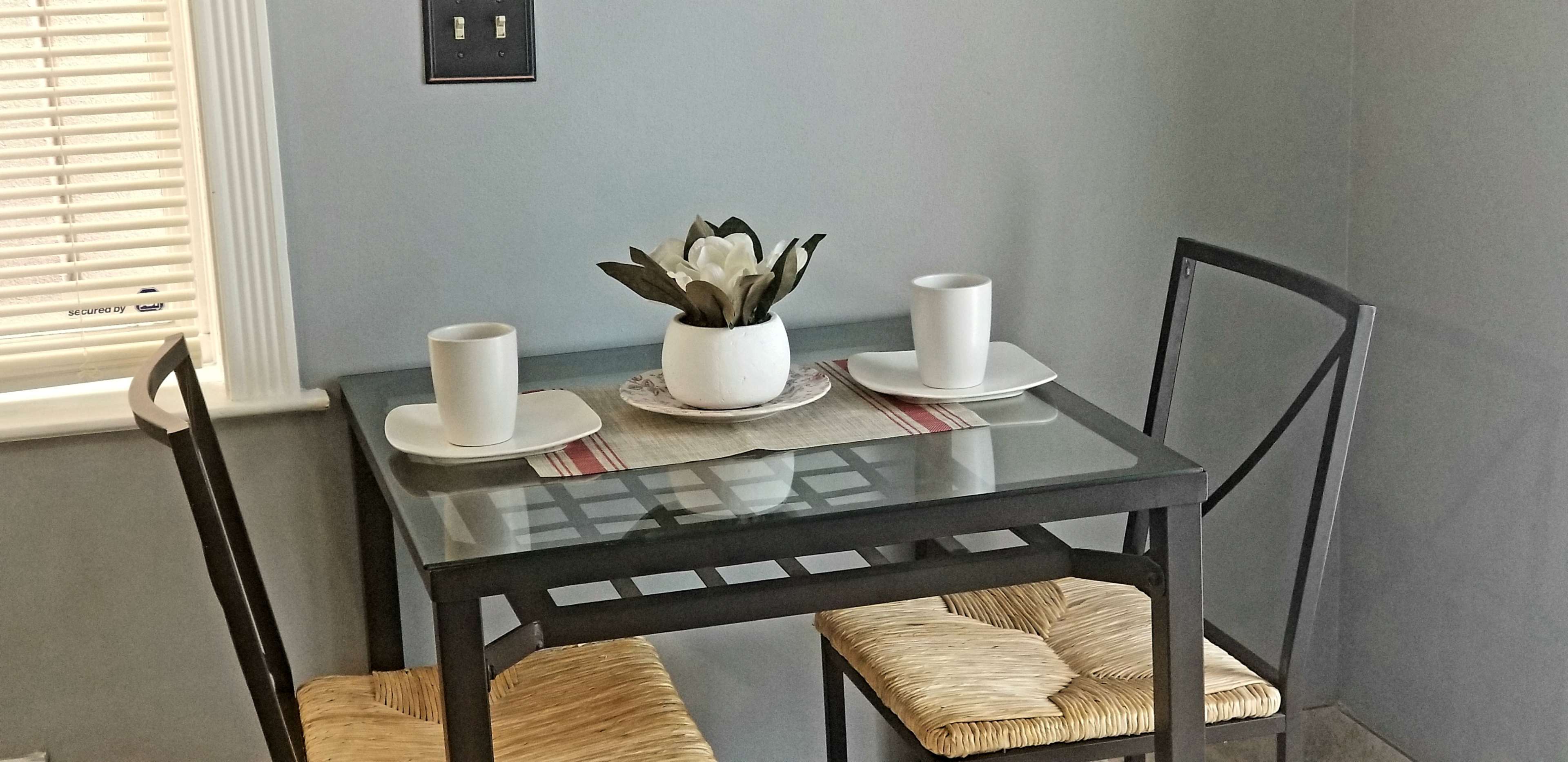 A small glass-top table with two chairs is set for two, featuring white mugs and a flower arrangement on a placemat.