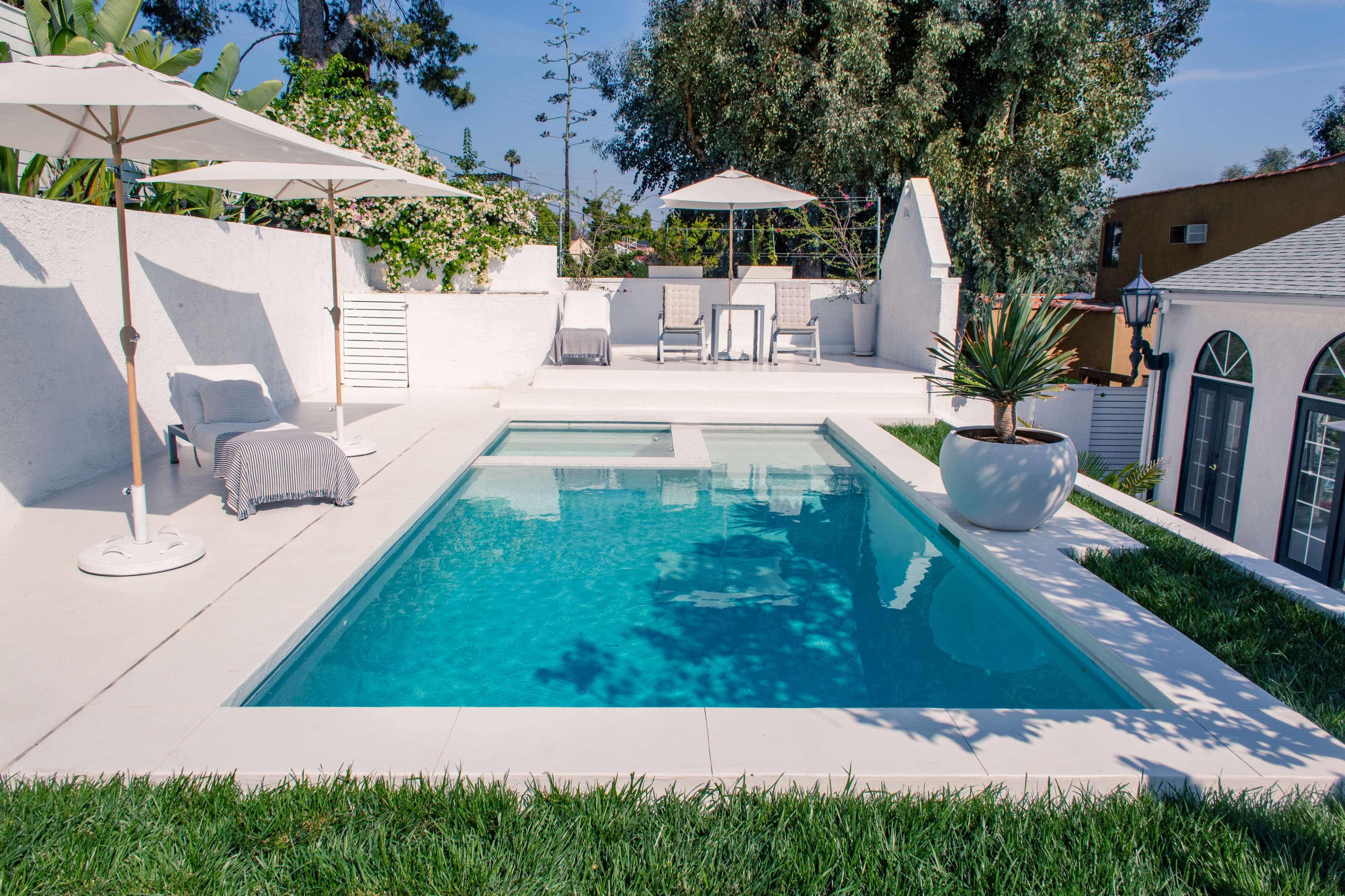 A modern pool area surrounded by white walls, featuring a clear swimming pool, lounge chairs, and shaded umbrellas.