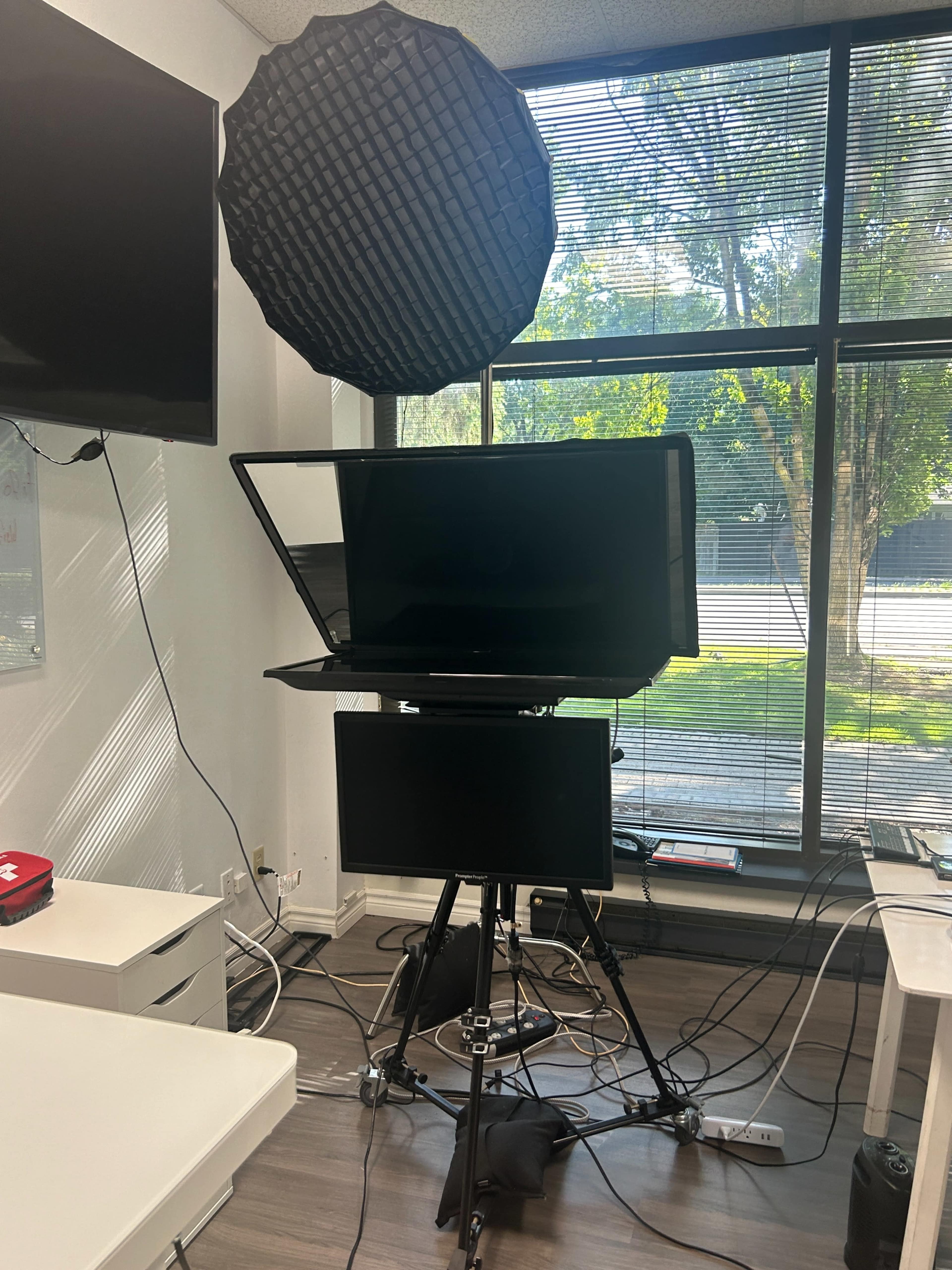 A setup featuring multiple monitors and lighting equipment stands in a room with large windows, revealing outside greenery.