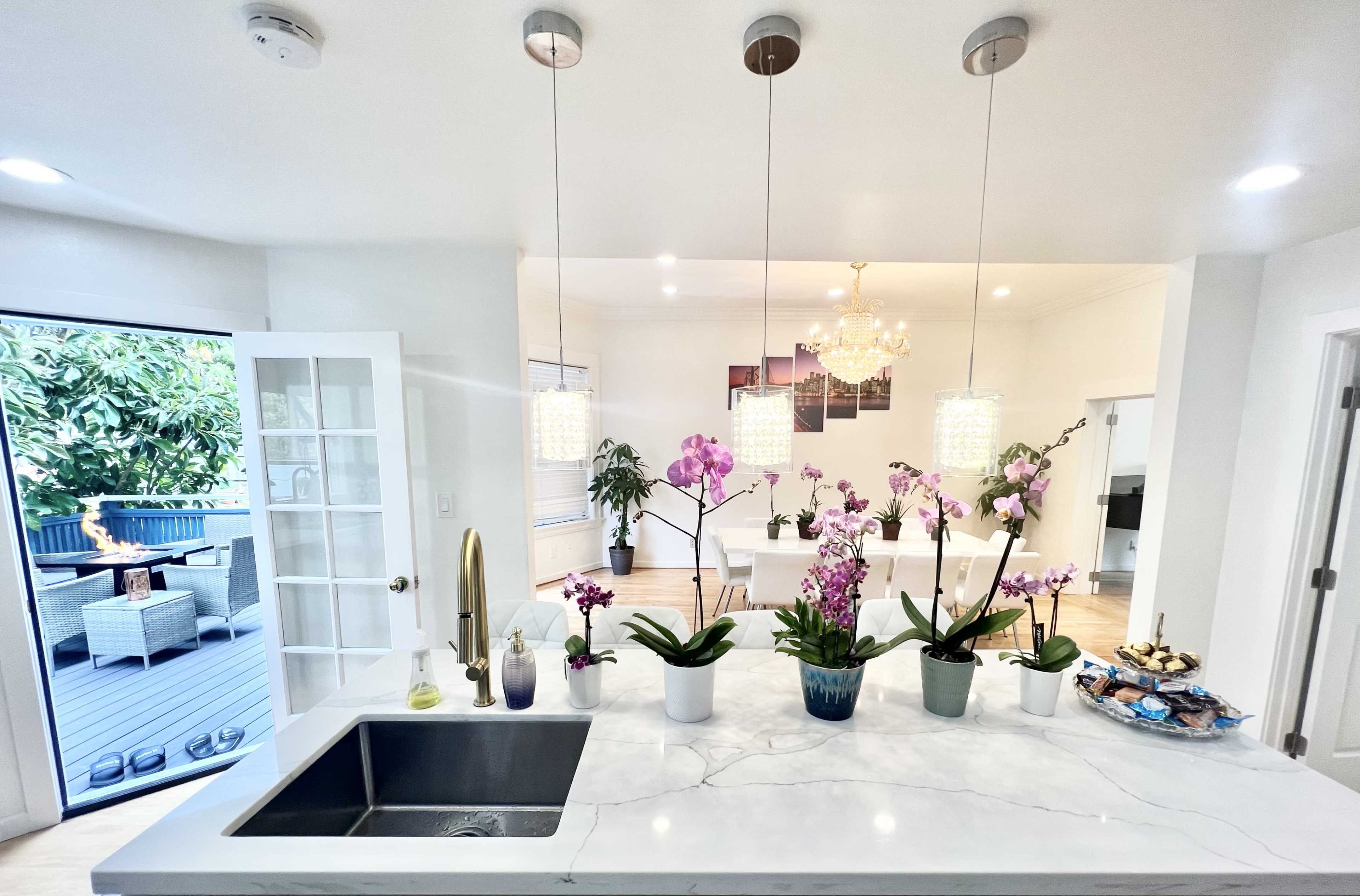 A kitchen features a marble countertop with a sink and several pots of orchids, illuminated by pendant lights, while a dining area is visible in the background.